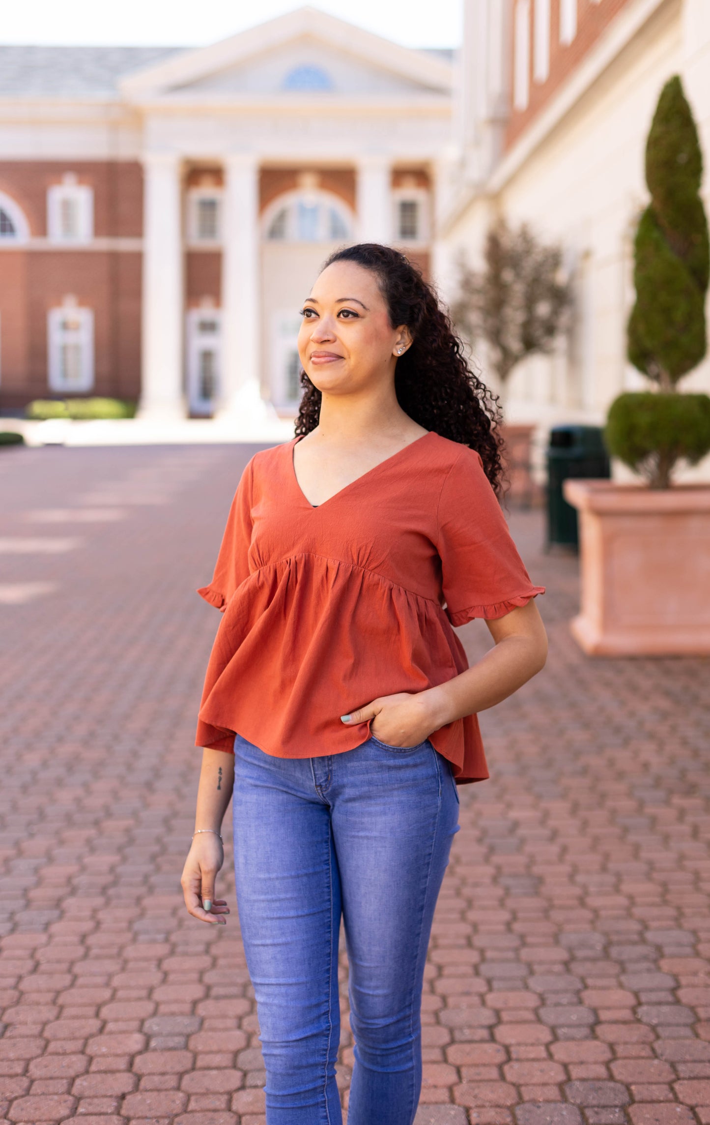 A woman in a rust-orange Dogwood Cloth Jenna Top and jeans walks on a paved path, with white columns and brick buildings in the background.