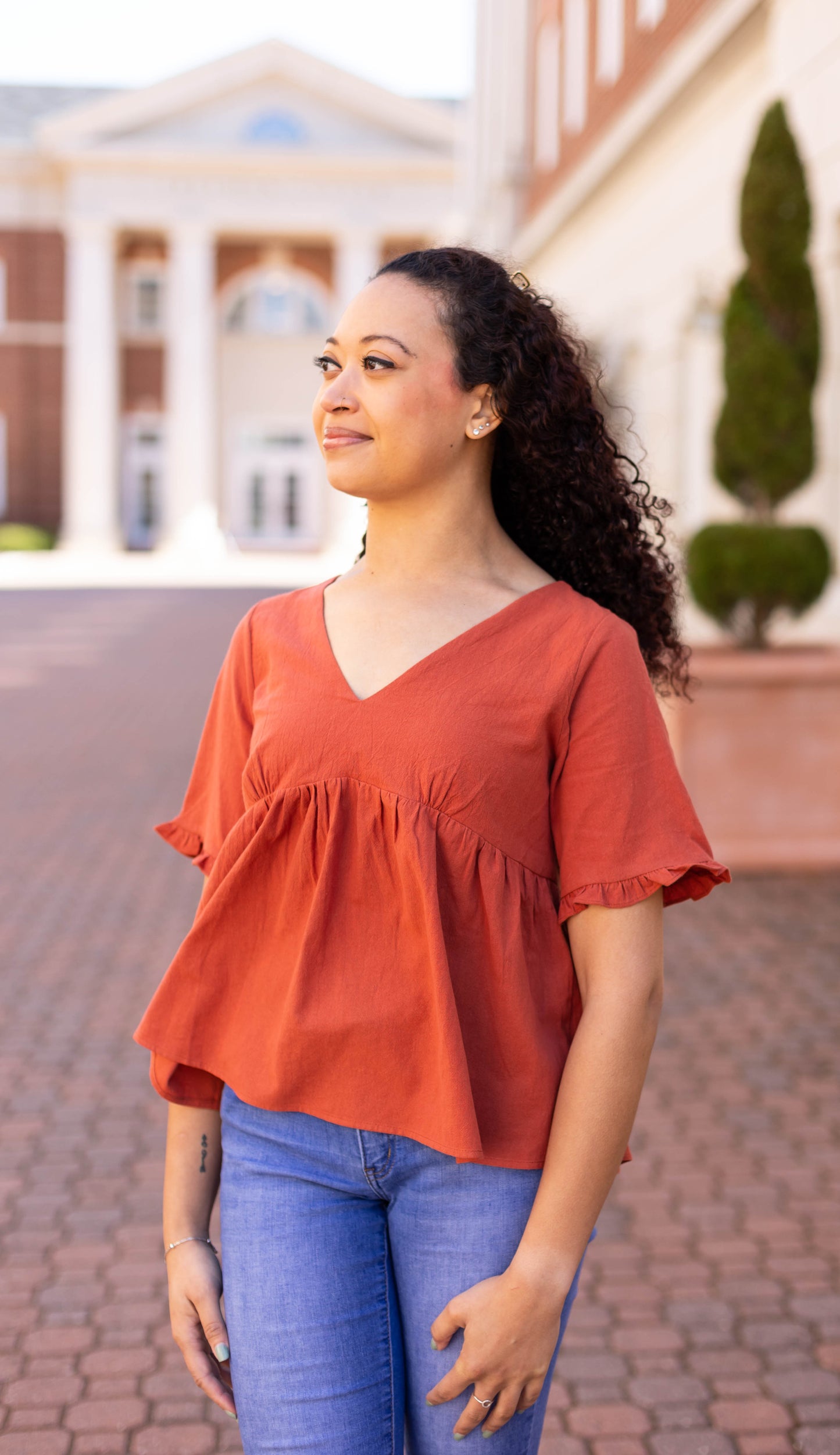 A woman wearing Dogwood Cloth's rust-orange Jenna Top and blue jeans stands on a cobblestone path, gazing to the side. Her curly hair bounces gently, framed by a building and lush greenery in the background.