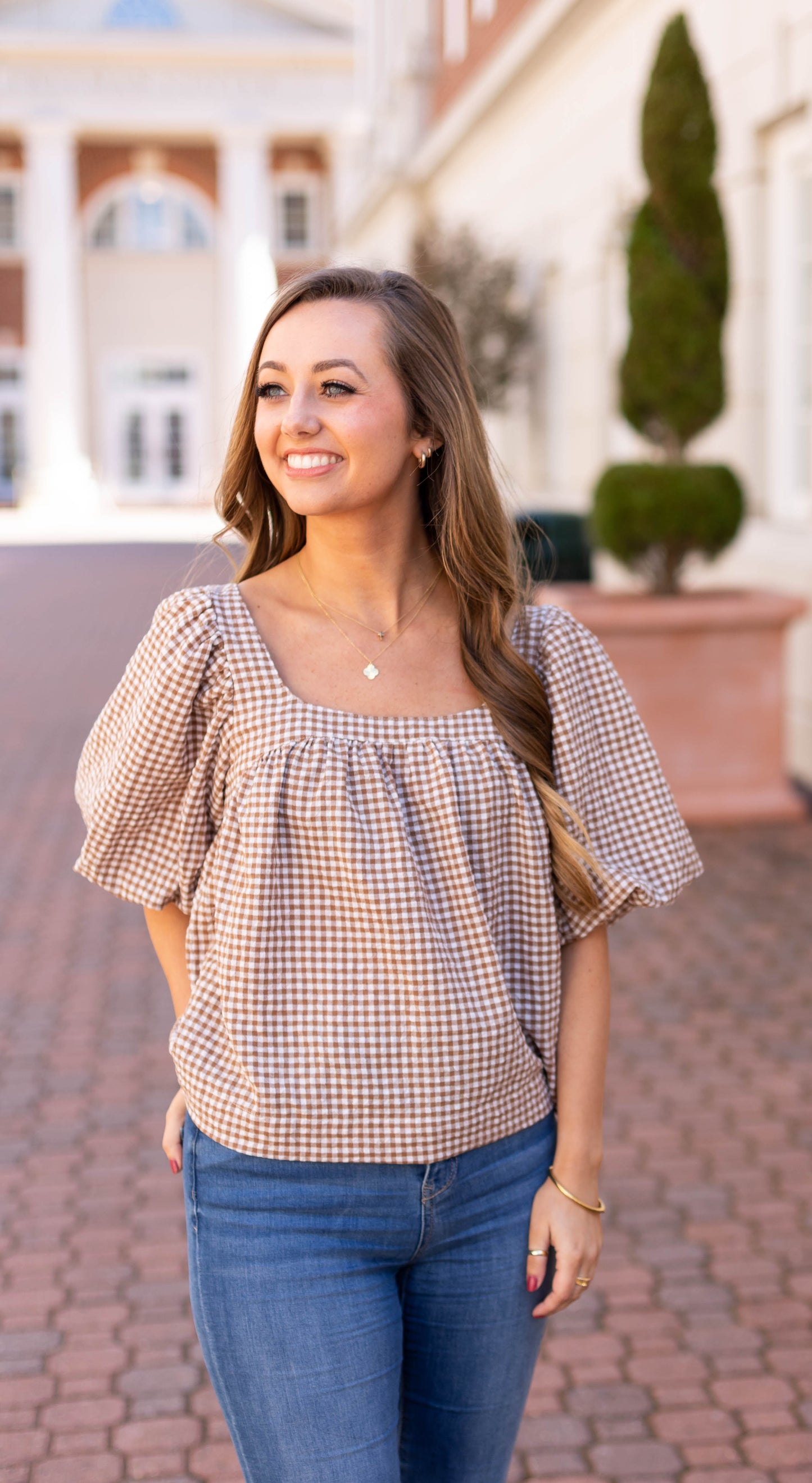 A woman radiates charm in her brown checkered Dogwood Cloth Bailey Gingham Top and jeans, standing on a brick path, smiling and gazing sideways, with background buildings.