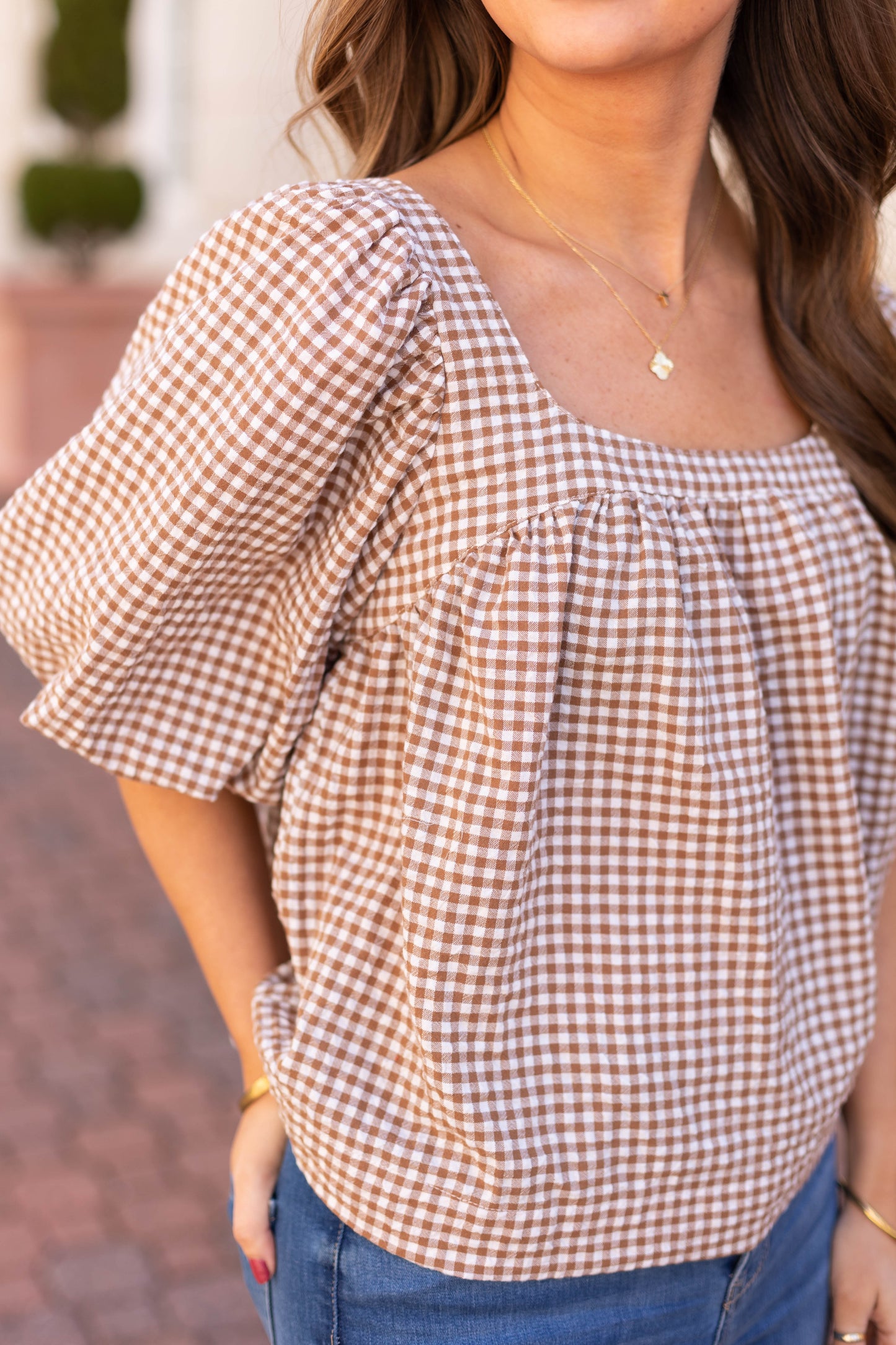 A person in a brown and white Bailey Gingham Top by Dogwood Cloth, featuring a babydoll fit, paired with blue jeans, exudes feminine charm while standing outdoors.