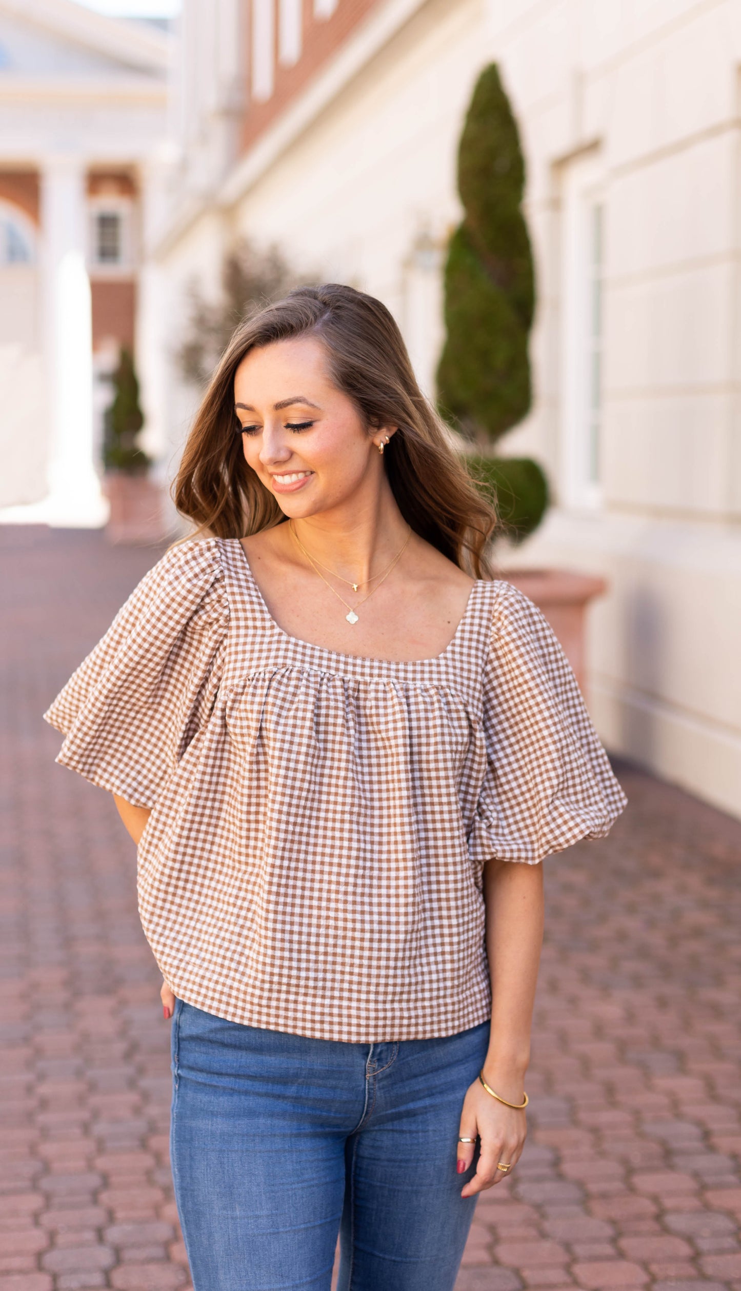 A woman with long hair stands outside on a brick pathway near a building with windows and neatly trimmed shrubs, exuding her feminine charm in a Dogwood Cloth Bailey Gingham Top.