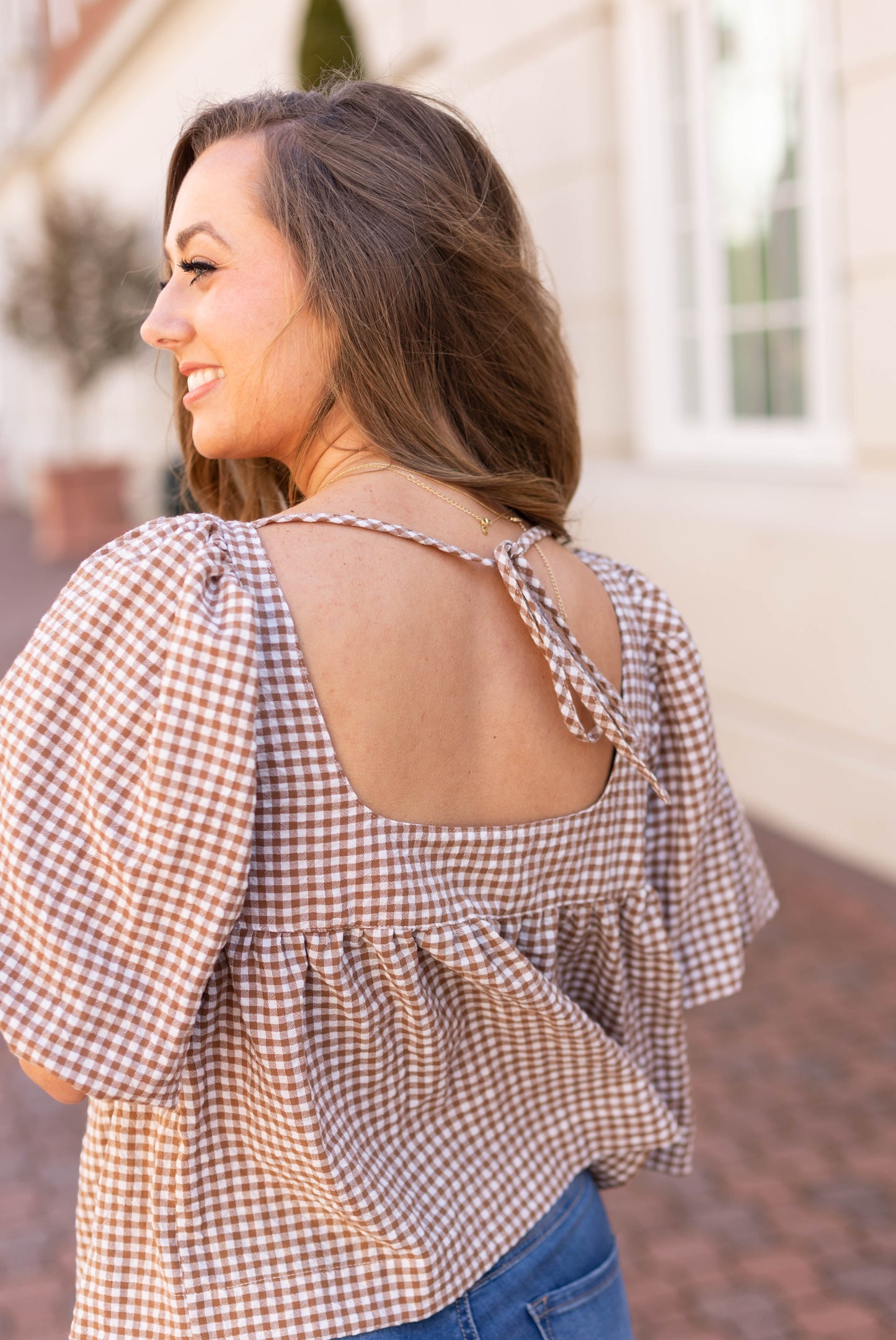 A woman in a Dogwood Cloth Bailey Gingham Top with a tie-back detail stands outdoors, exuding feminine charm as she smiles and looks to the side.