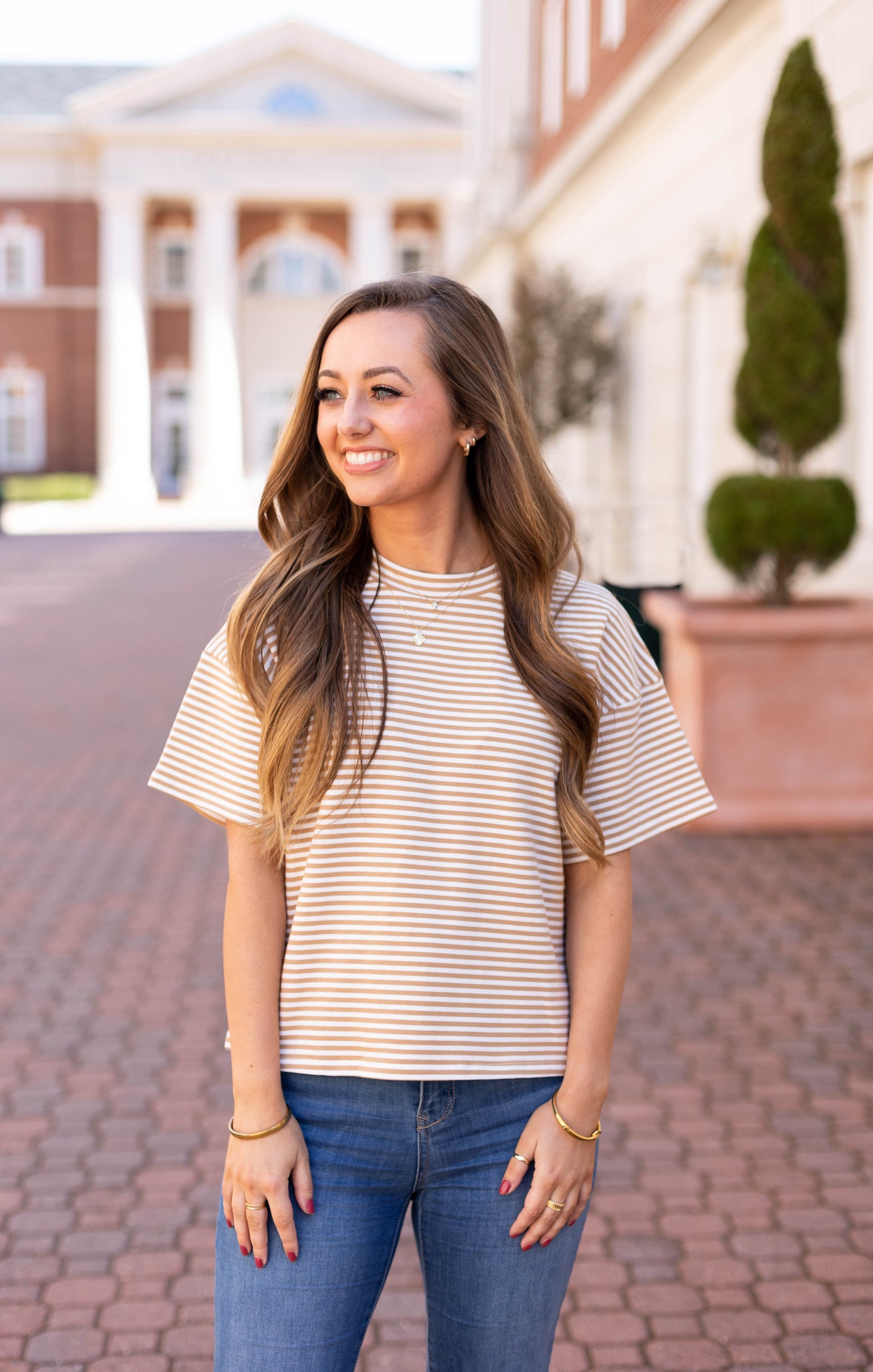 A woman in a Dogwood Cloth Delaney Striped Tee and jeans smiles on a brick path, embodying casual comfort against a building with columns, capturing everyday wardrobe essence.
