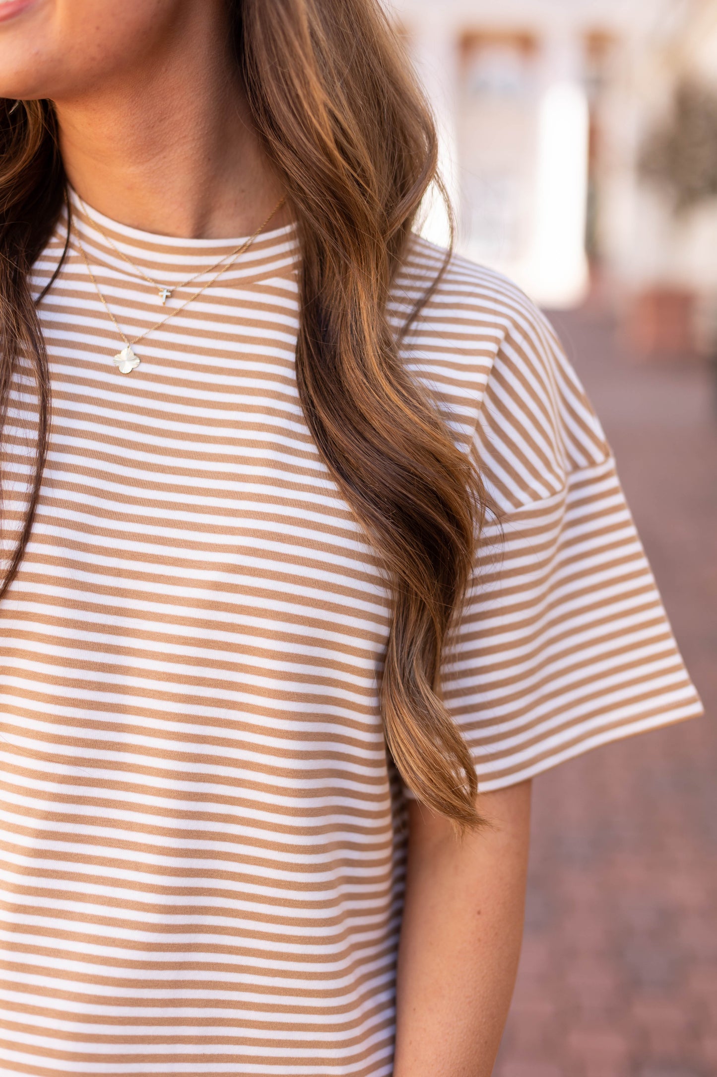 Person with long wavy hair in a beige and white Dogwood Cloth Delaney Striped Tee paired with a necklace, capturing casual comfort against a blurred backdrop.