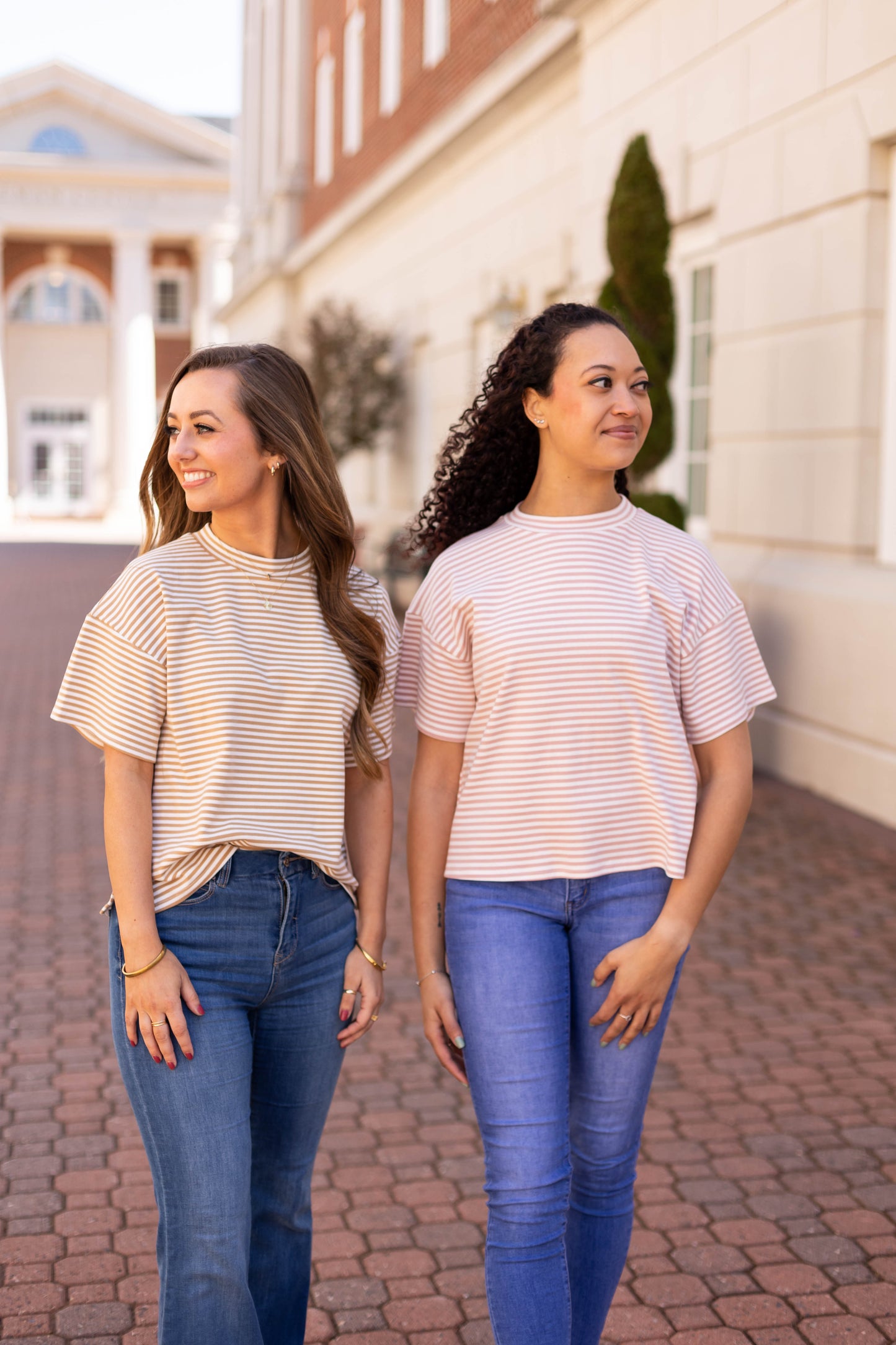 Two women in Dogwood Cloth's Delaney Striped Tees and jeans stand on a brick pathway in an urban setting, embodying casual comfort and everyday essentials with buildings framing their backdrop.