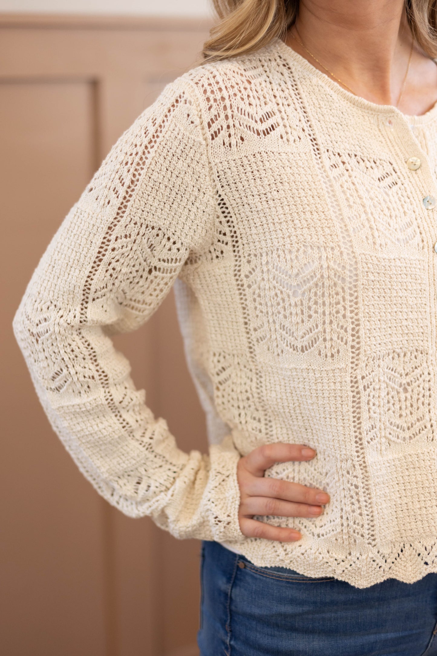 A person wearing the Claire Lace Sweater by Dogwood Cloth, a cream long-sleeve cotton-polyester top with geometric crochet patterns, and blue jeans. Hand wash cold and cool iron recommended for care.
