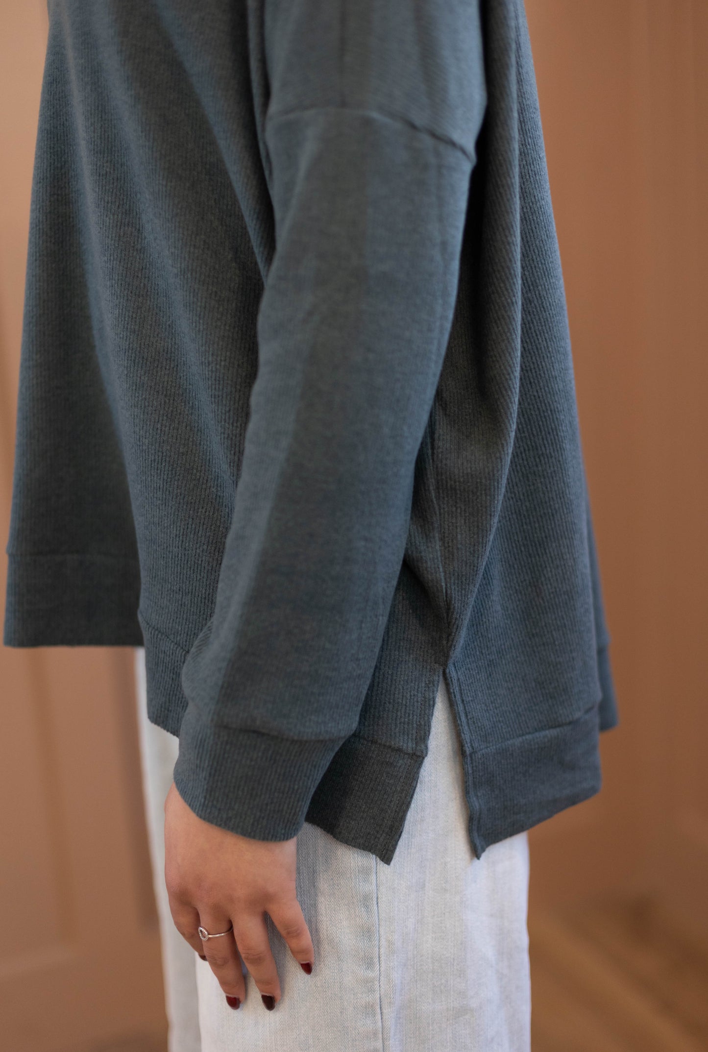 A person wearing the Chandler Sage Top by Dogwood Cloth, a cozy blue women’s sweater with a side slit, paired with light-colored pants and standing indoors with their arm relaxed at their side.