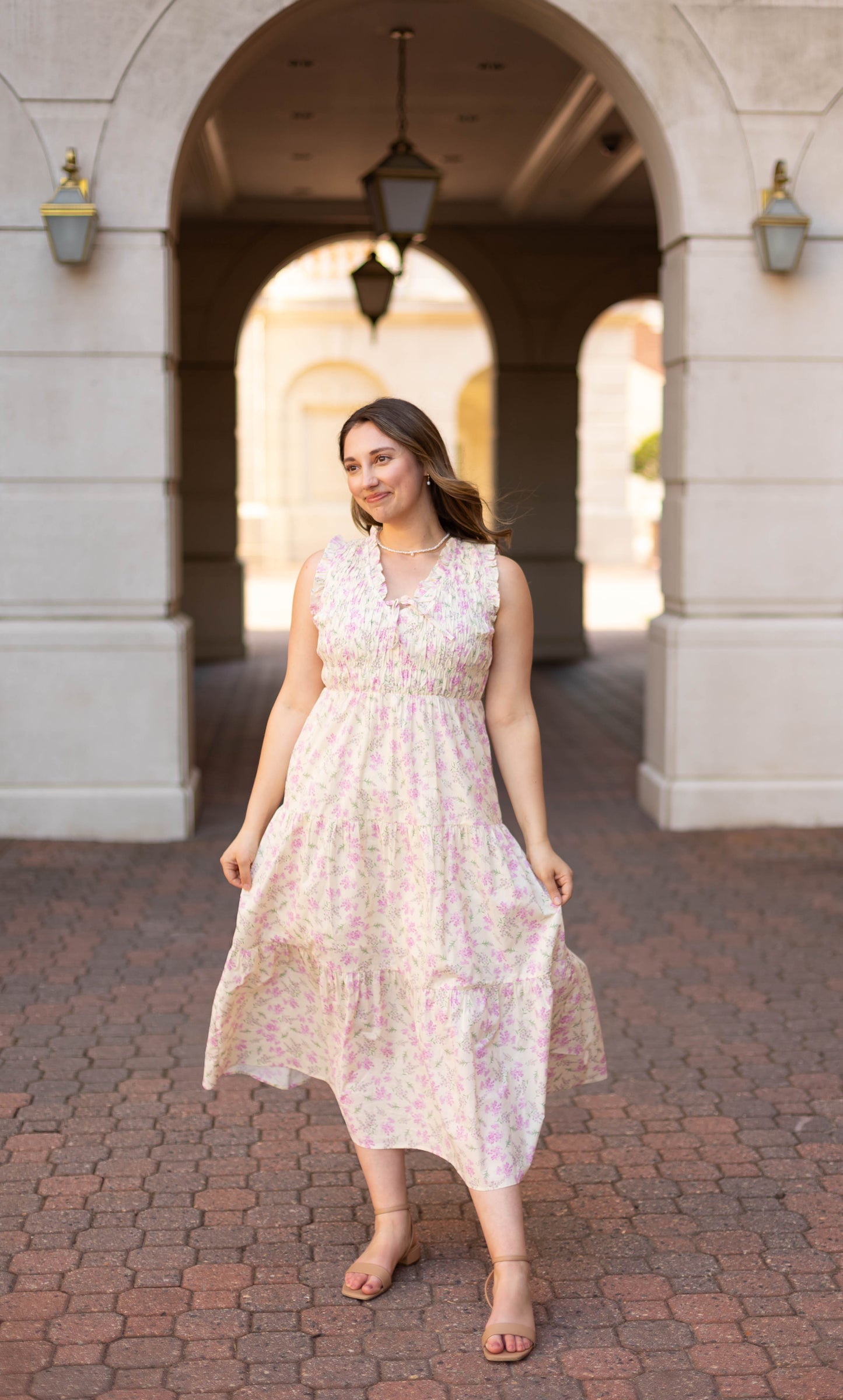 A woman in the Dogwood Cloth Sophia Floral Midi stands on a brick pathway under an archway, facing forward with a slight smile.