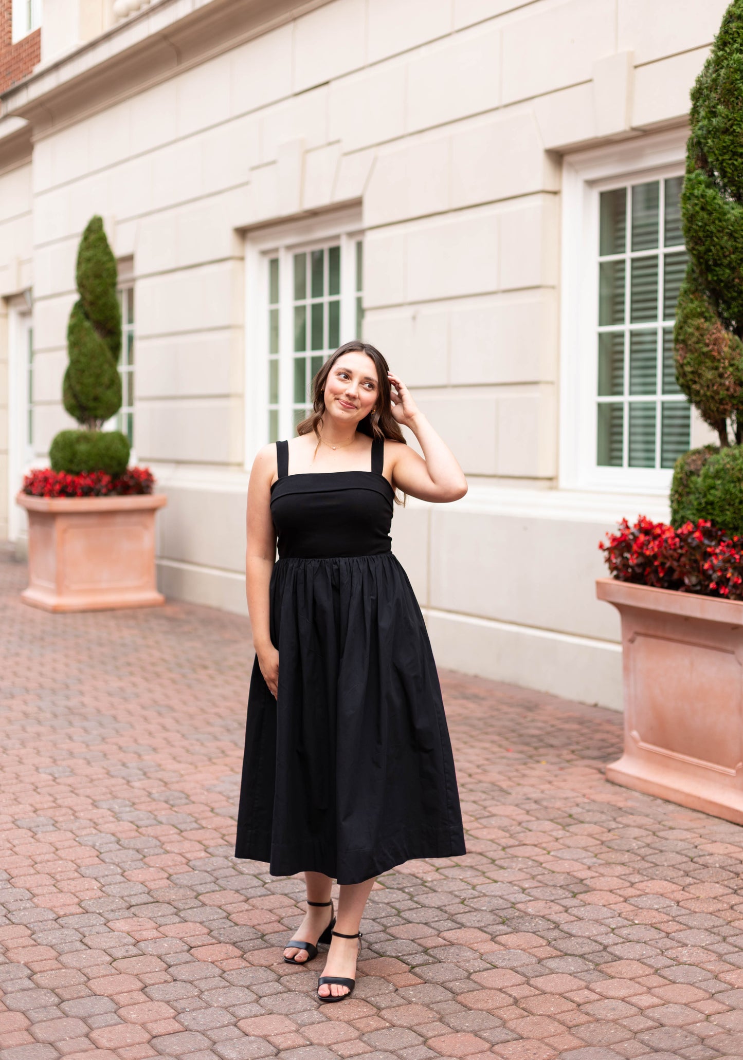 A woman wearing the Dogwood Cloth Evelyn Classic Midi, featuring an A-line silhouette, stands on a brick walkway before a beige building with white-trimmed windows and large planters of red flowers and sculpted shrubs.