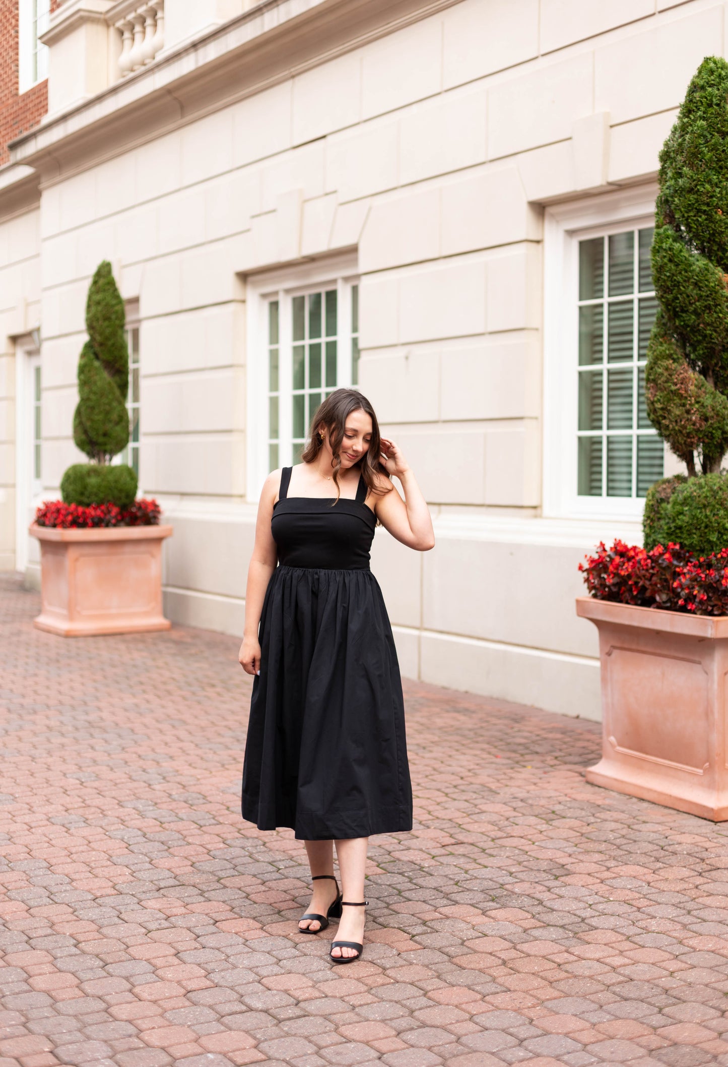 A woman in Dogwood Cloth's The Evelyn Classic Midi, featuring an A-line silhouette, pairs it with black sandals as she stands on a brick walkway before a beige building adorned with windows and large potted plants.