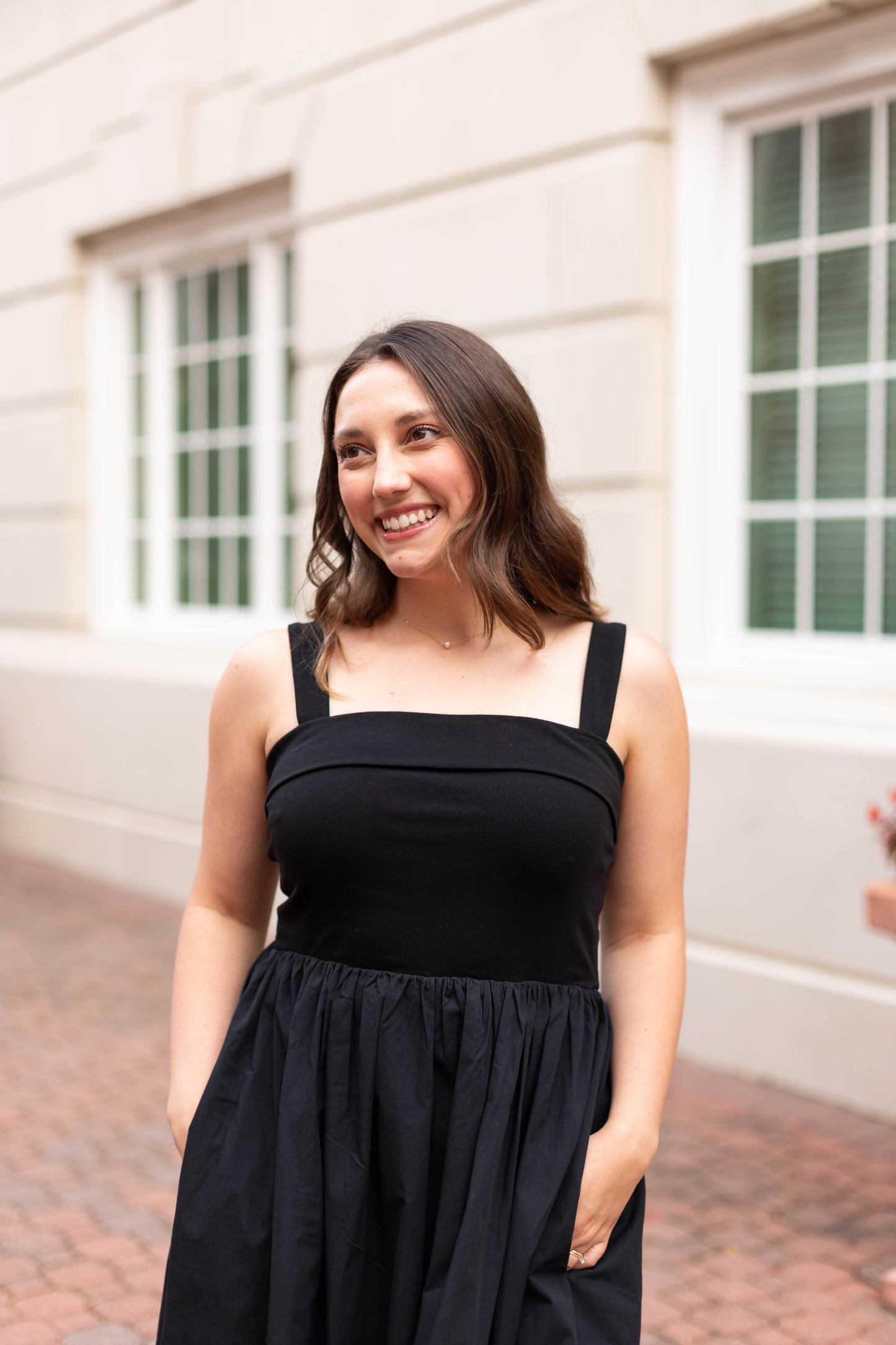 A woman wearing Dogwood Cloth’s The Evelyn Classic Midi stands outside a light-colored building with large windows, smiling and looking to the side.