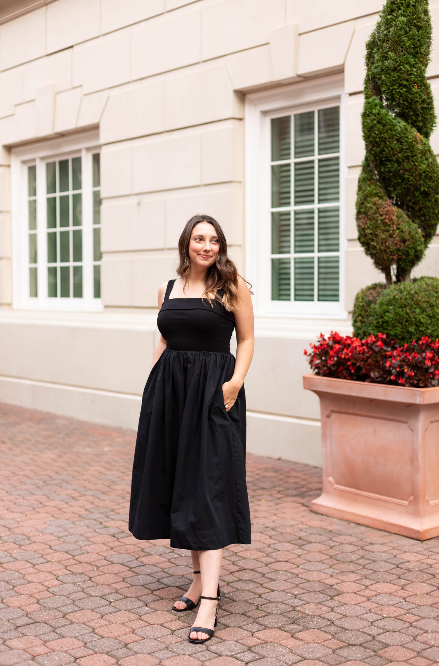 A woman wears Dogwood Cloth’s The Evelyn Classic Midi, an A-line black dress, paired with black sandals as she stands on a brick sidewalk near a beige building with two windows and a large potted plant.