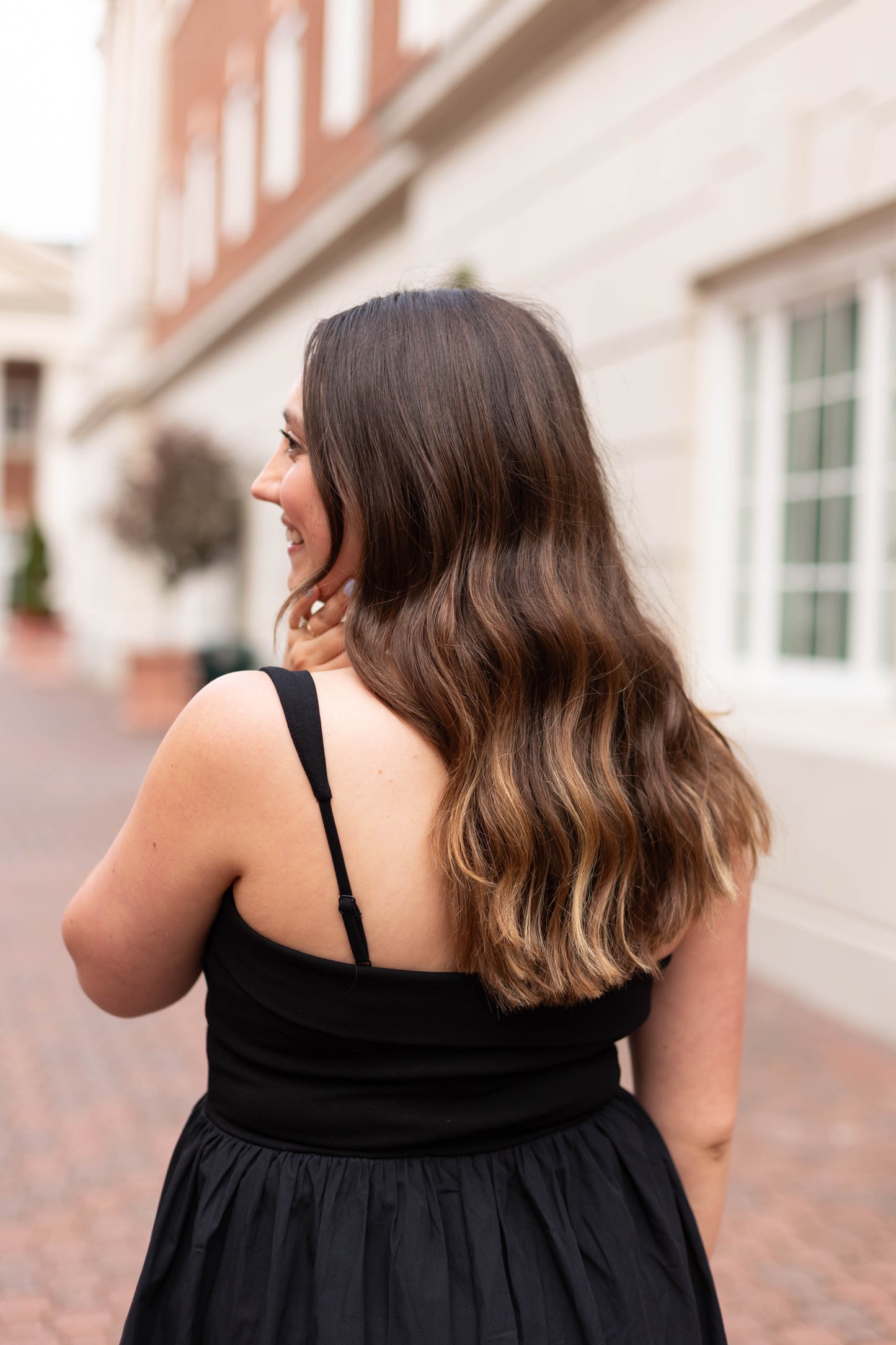 A woman with long wavy brown hair, wearing The Evelyn Classic Midi by Dogwood Cloth, stands outdoors on a brick walkway near white buildings, facing away from the camera.