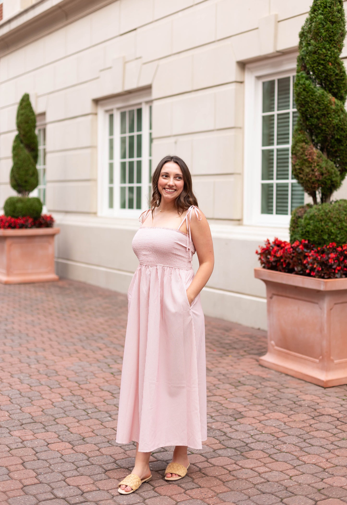 A woman wearing the Dogwood Cloth Summer Pin-Stripe Maxi, a light pink maxi dress with a smocked bodice, stands on a brick walkway in front of large windows and decorative plants.