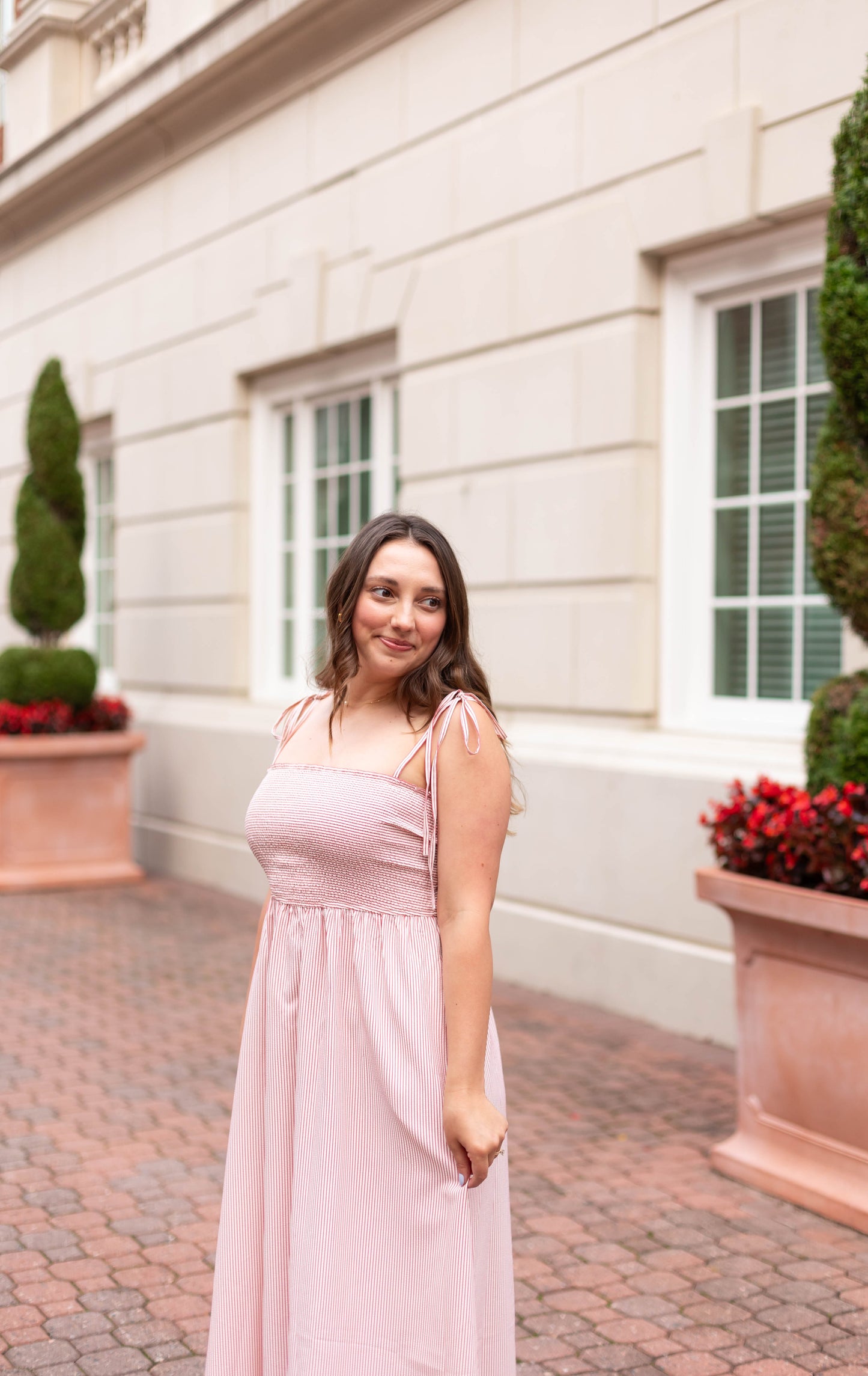 A woman wearing the Dogwood Cloth Summer Pin-Stripe Maxi stands on a brick walkway in front of a beige building with large windows and potted plants.
