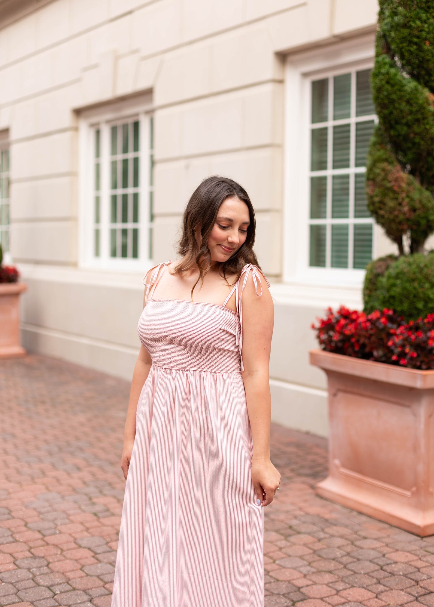 A woman wearing the Dogwood Cloth Summer Pin-Stripe Maxi stands on a brick path near a building with large windows and potted plants.