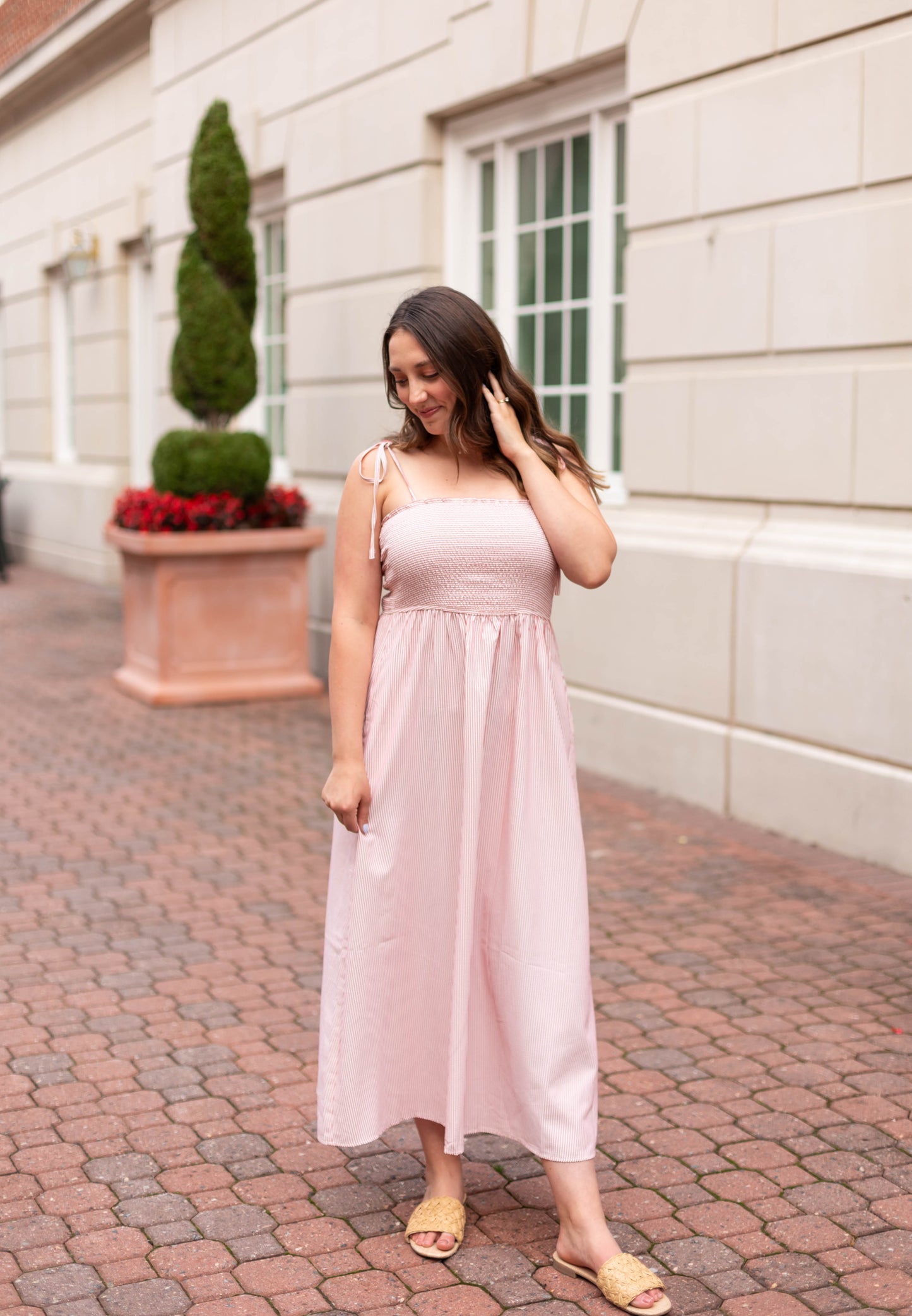 A woman wearing the Dogwood Cloth Summer Pin-Stripe Maxi with a smocked bodice and sandals stands on a brick walkway near large windows and a decorative planter.