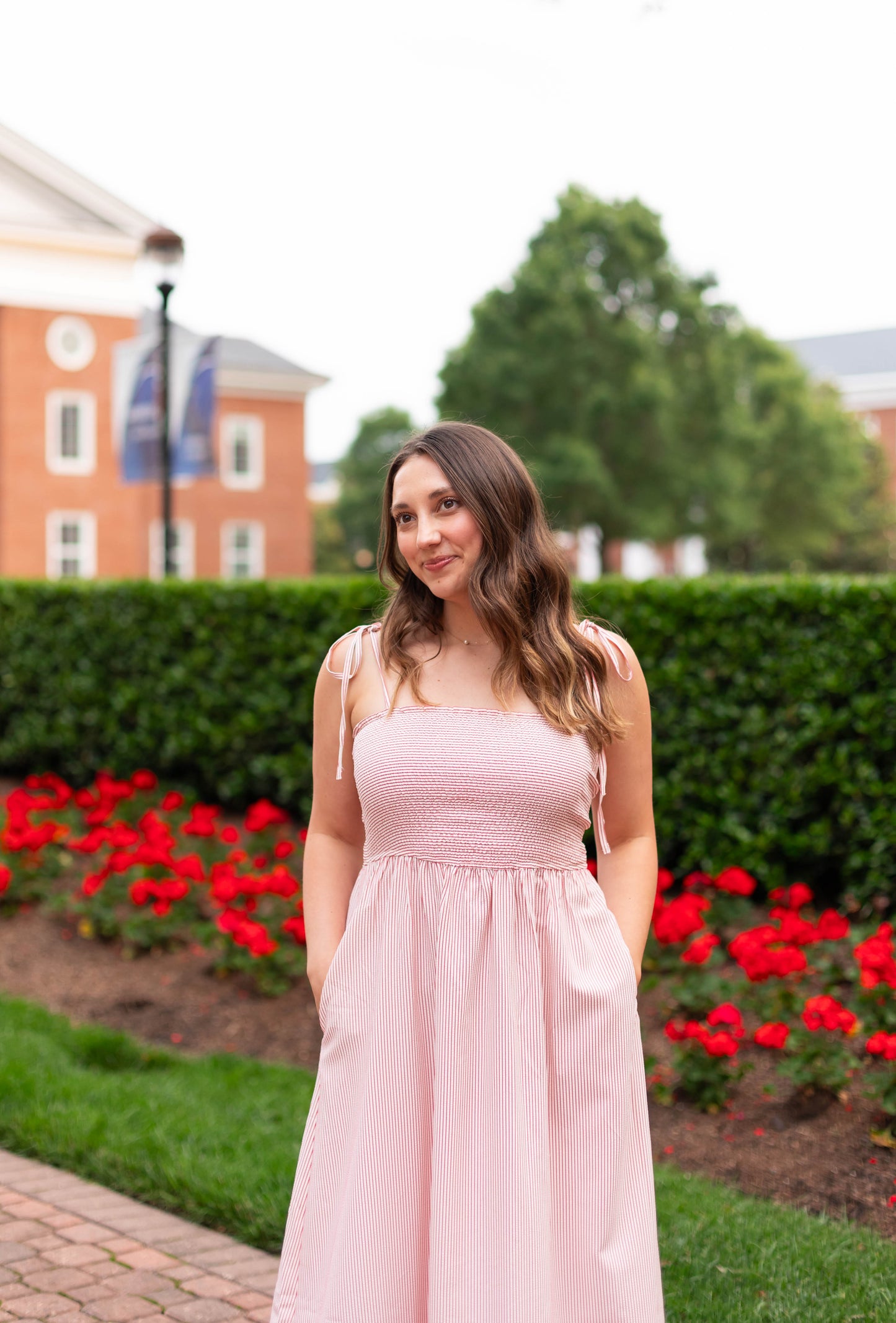 A woman in the Dogwood Cloth Summer Pin-Stripe Maxi with spaghetti straps stands outdoors on a brick path, surrounded by red flowers, green hedges, and campus buildings in the background.