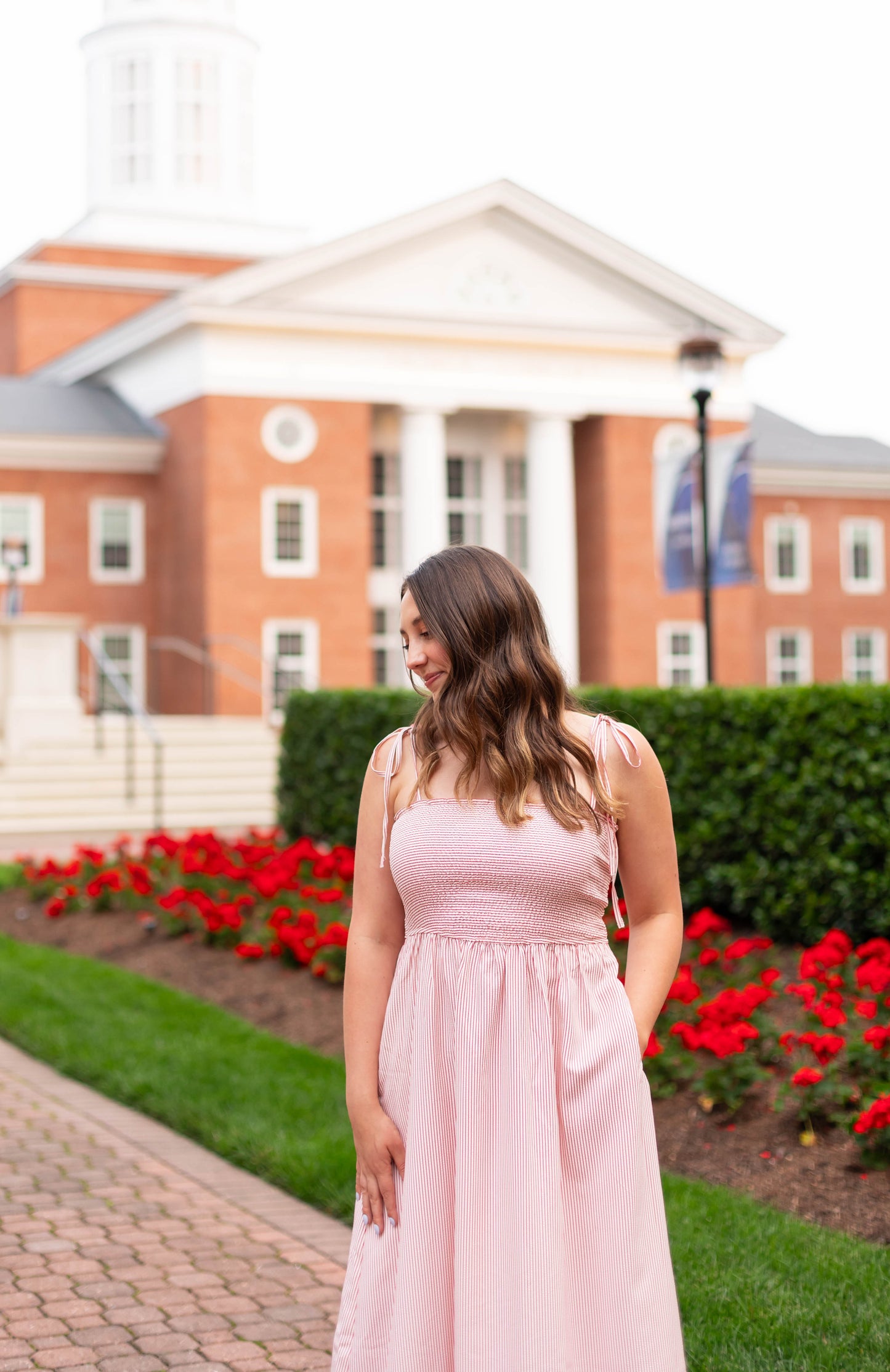 A woman wears the Dogwood Cloth Summer Pin-Stripe Maxi while standing on a brick path near a red brick building with white columns, surrounded by red flowers and greenery.
