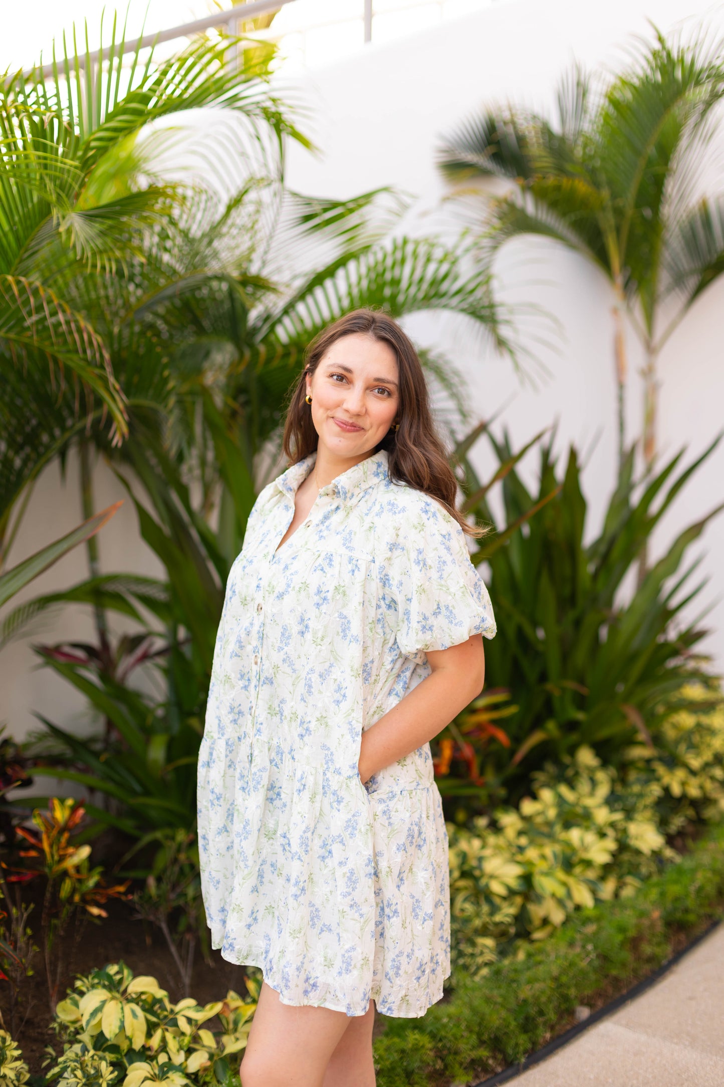 A woman wears The Margot Floral Dress by Dogwood Cloth, featuring puff sleeves, as she stands outdoors among green plants and tropical foliage, gazing at the camera with a gentle smile.