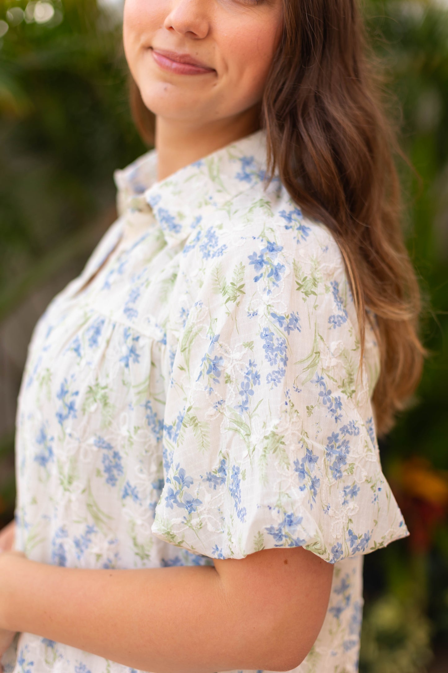A woman with long brown hair stands outdoors wearing Dogwood Cloth’s Margot Floral Dress, a light short-sleeve blouse with blue and green floral patterns and delicate embroidered details.