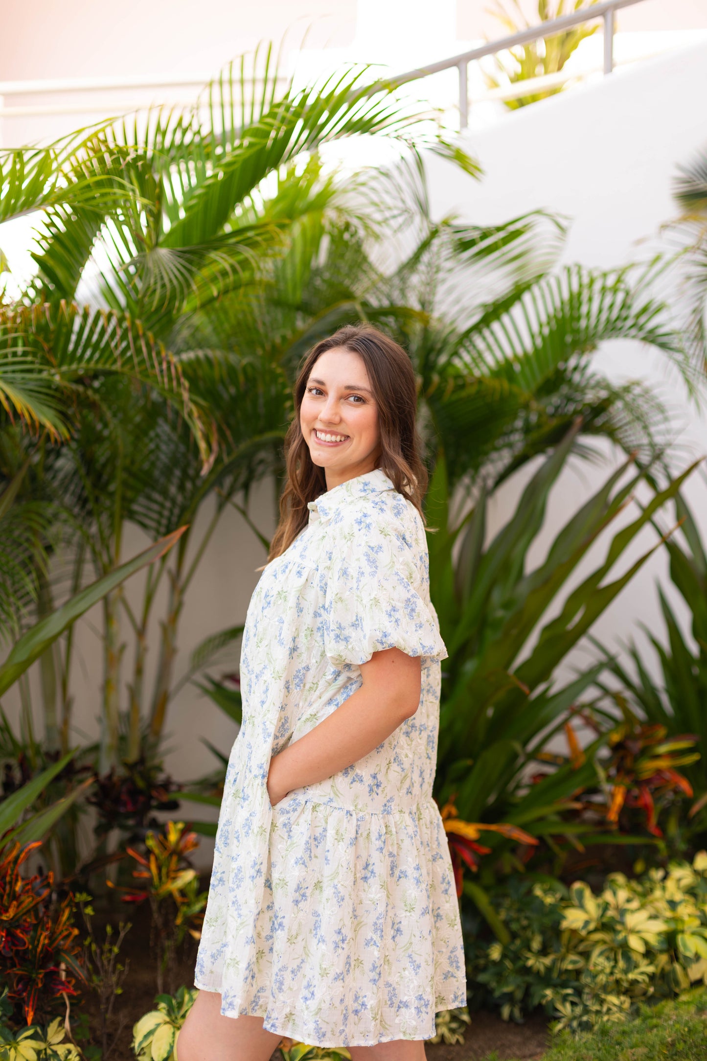 A young woman wearing The Margot Floral Dress by Dogwood Cloth, featuring puff sleeves, smiles at the camera while standing outdoors among green plants.