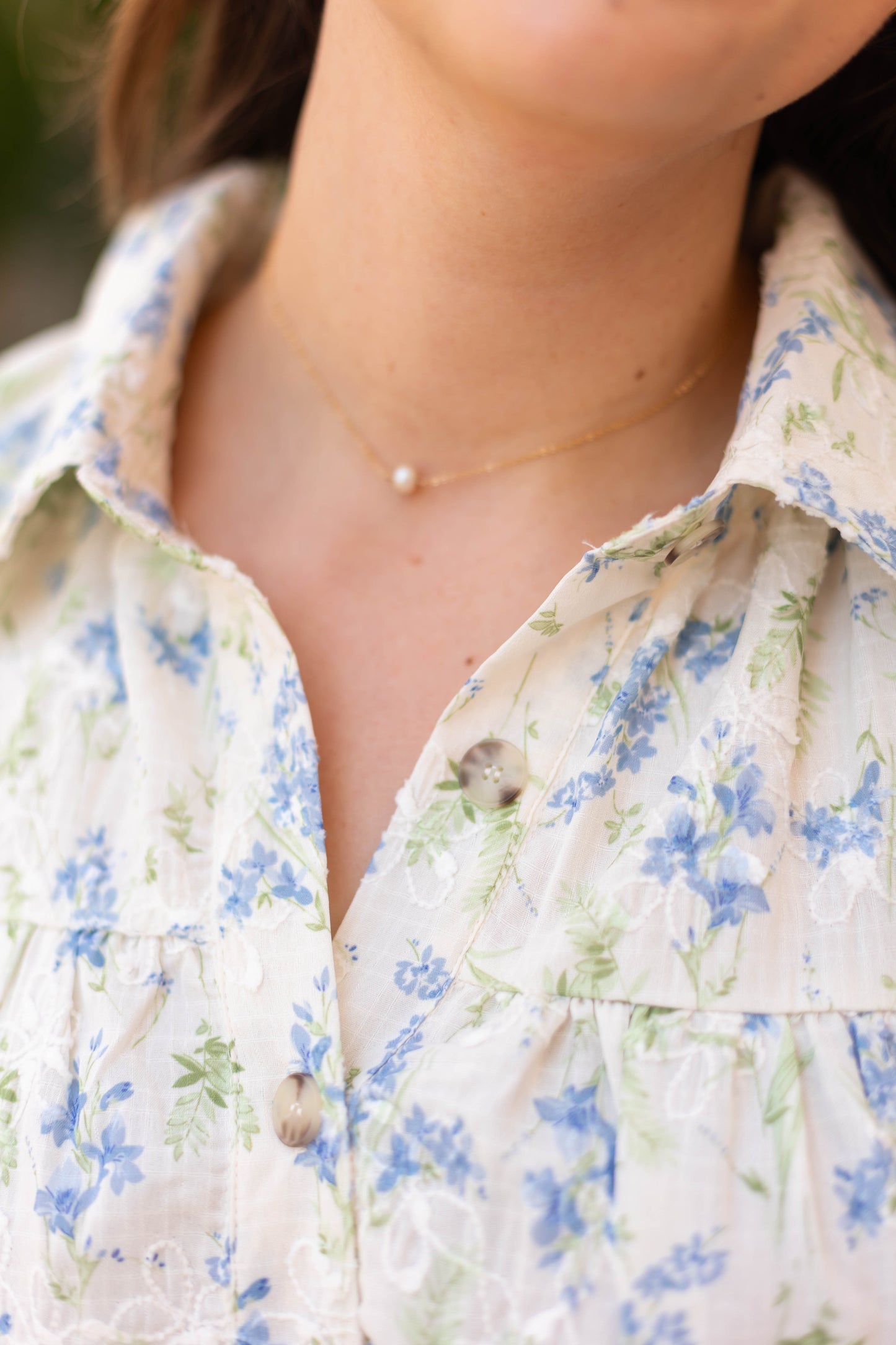 Close-up of a person wearing Dogwood Cloth's Margot Floral Dress, adorned with blue and green flowers, delicate embroidered details, and paired with a gold necklace featuring a small pearl pendant.
