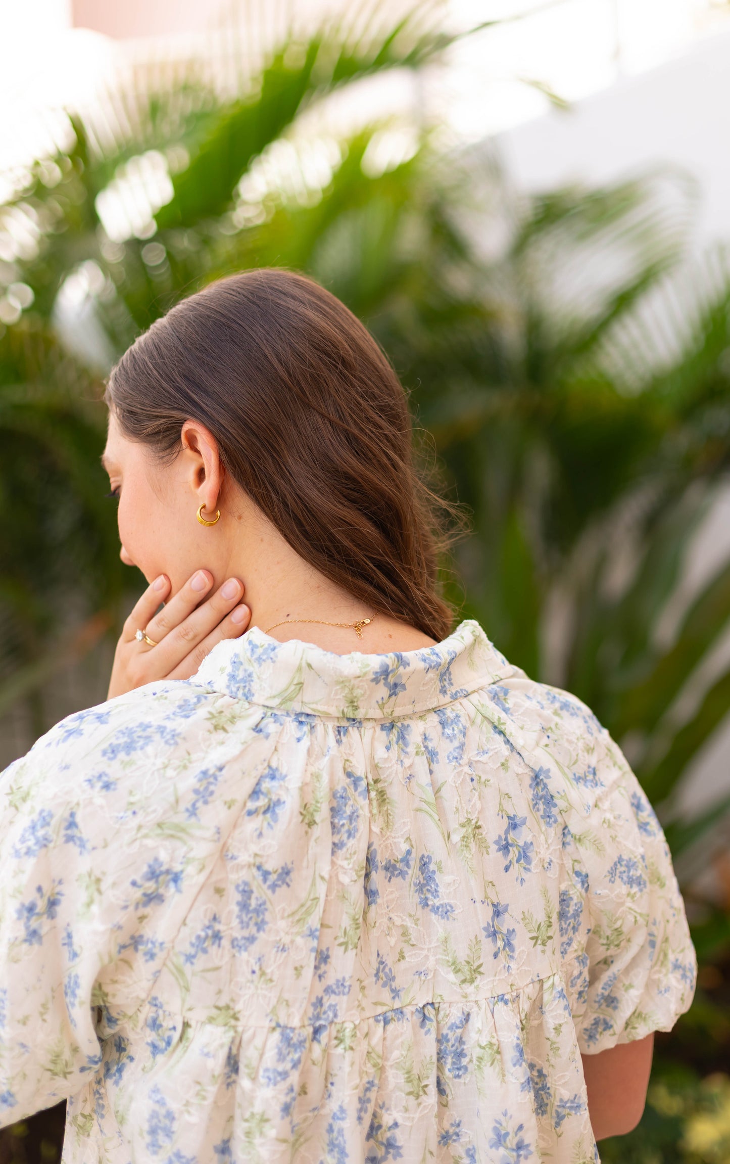 A woman with long brown hair, wearing Dogwood Cloth’s Margot Floral Dress with puff sleeves and gold earrings, stands outdoors facing away, touching her neck. Lush green plants fill the background.