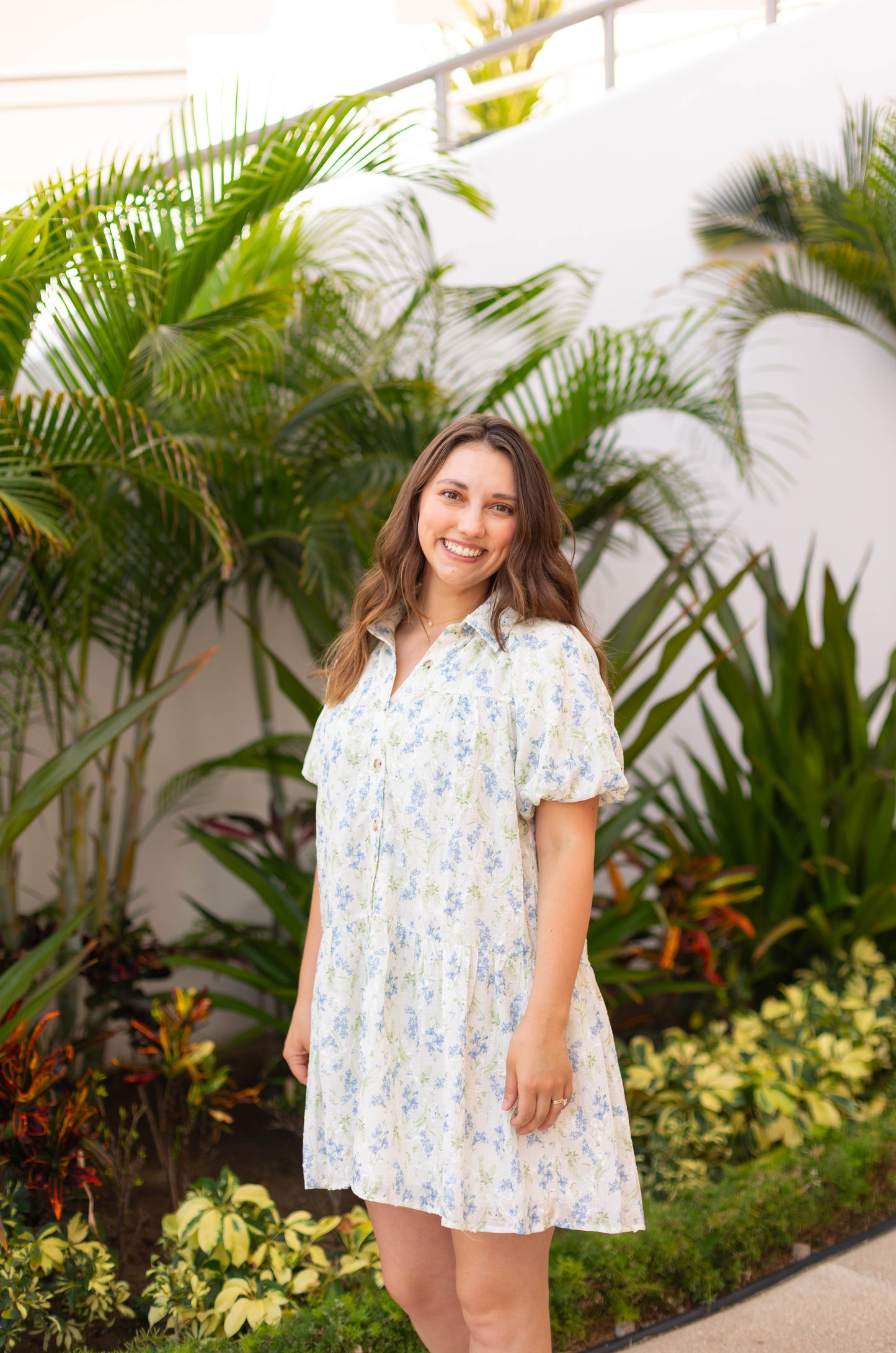 A woman in The Margot Floral Dress by Dogwood Cloth—featuring light blue floral fabric and puff sleeves—smiles in front of tropical greenery.
