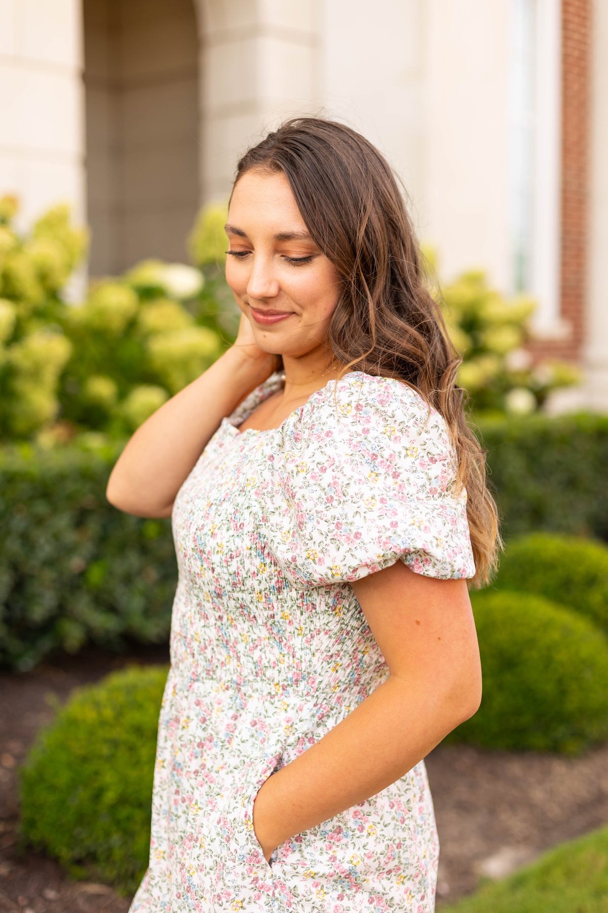 A woman with long brown hair wears the Dogwood Cloth Meadow Tiered Maxi, standing outdoors in front of green bushes and looking down with a slight smile.