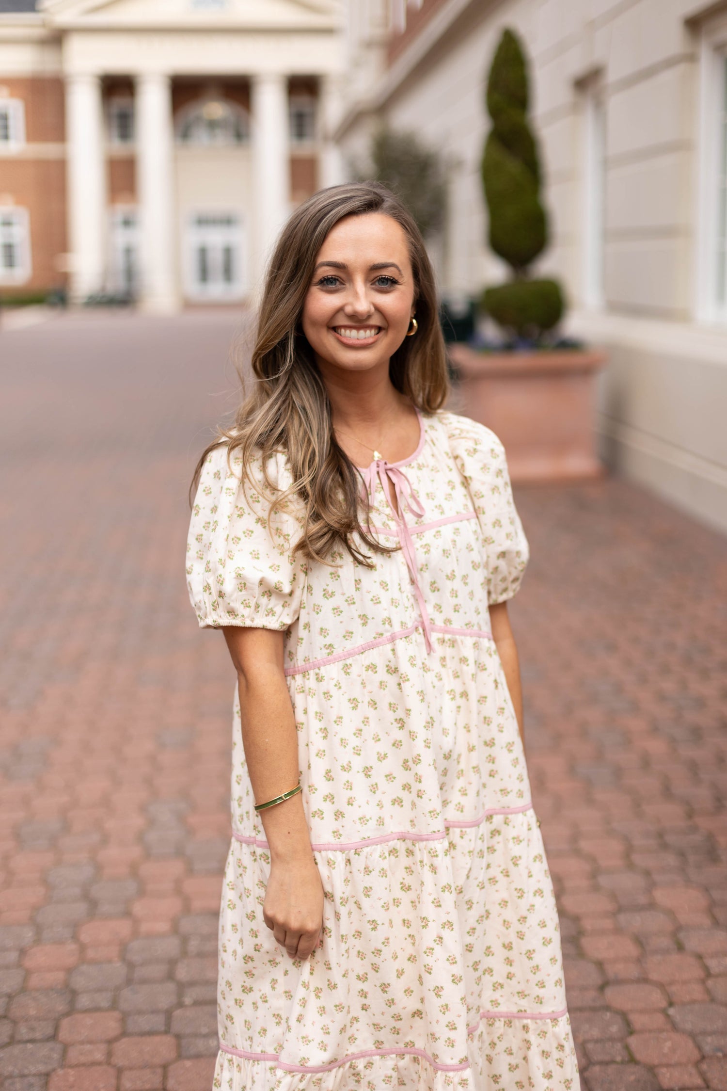 A woman in a light floral dress stands on a brick walkway in front of a building with columns and trimmed greenery, smiling at the camera.