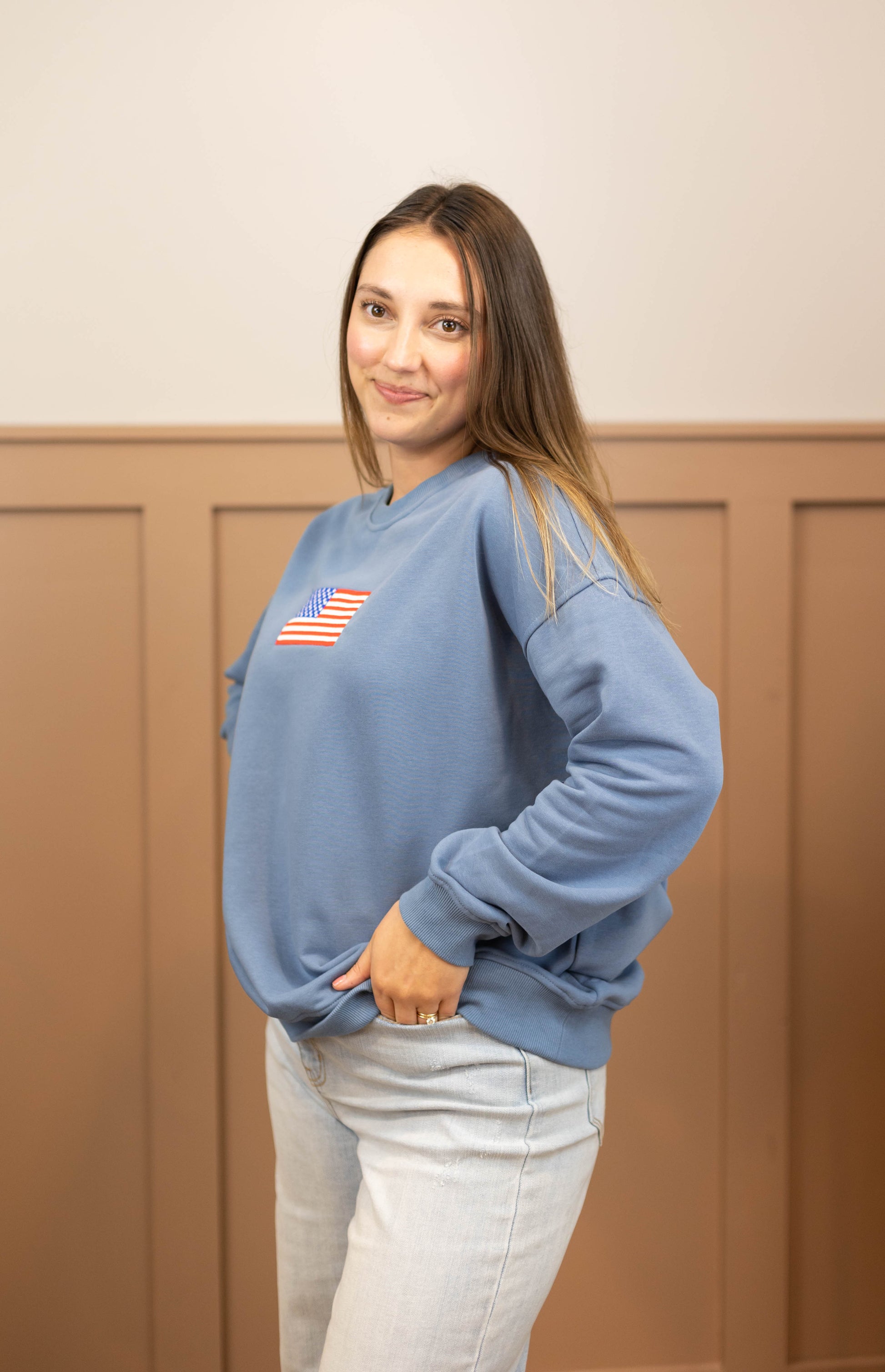 A person wearing a denim blue Liberty Crewneck Pullover from Dogwood Cloth, showcasing an American flag design, stands with their hands in pockets against a beige paneled wall.