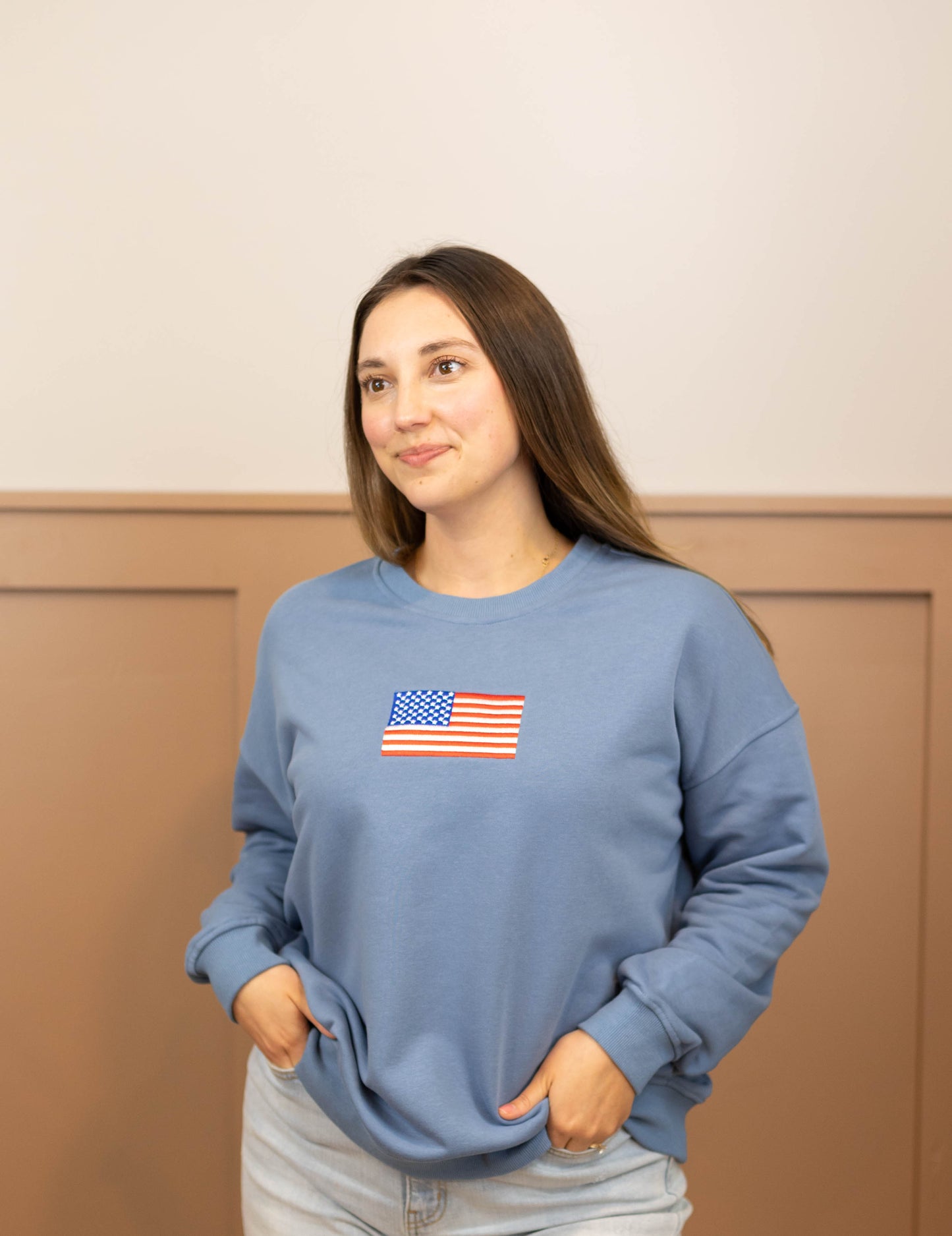 A person wearing a denim blue Liberty Crewneck Pullover from Dogwood Cloth, featuring an American flag design, stands against a beige and brown wall.