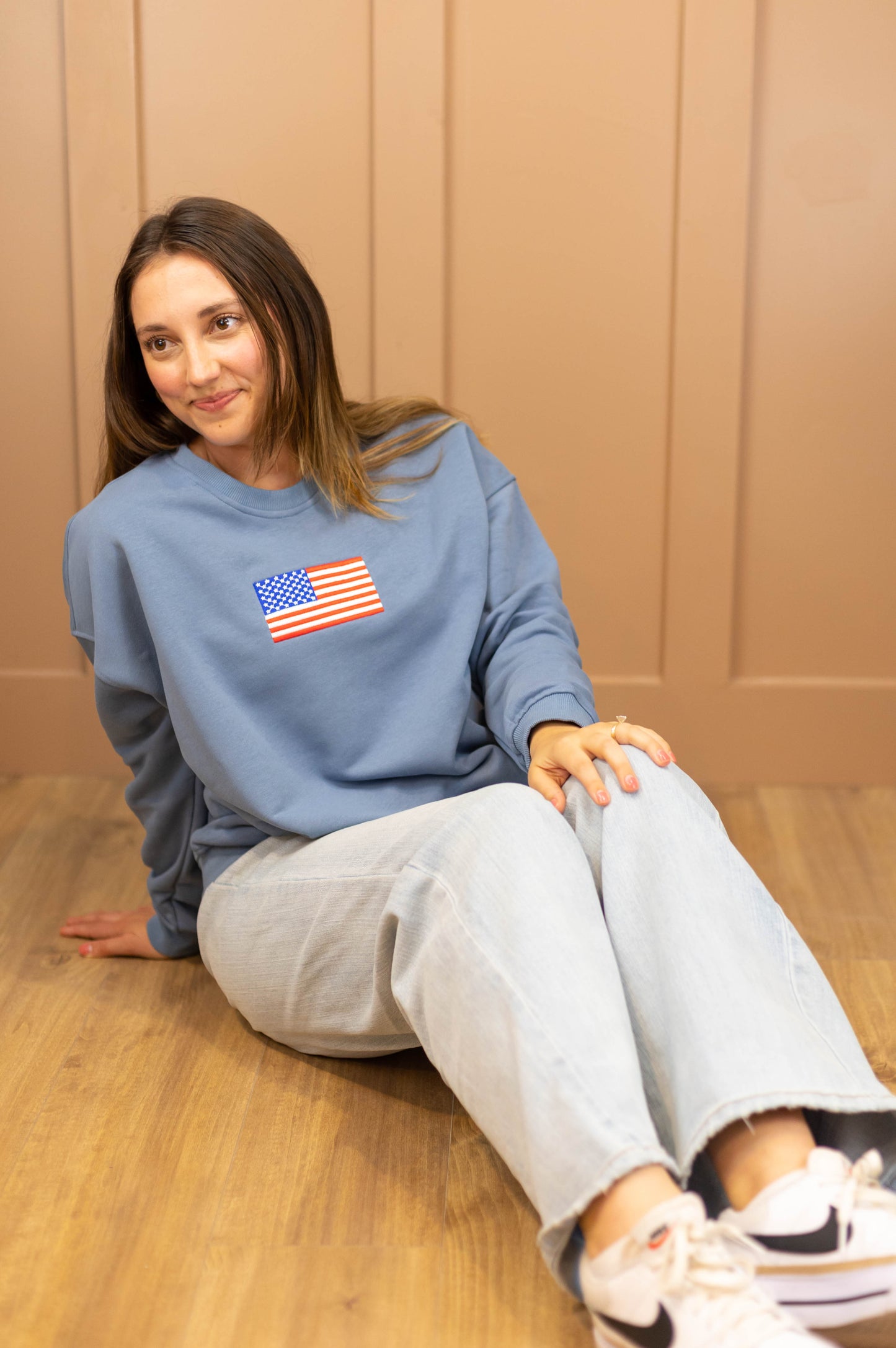 A person with long hair sits on a wooden floor, wearing a denim blue Liberty Crewneck Pullover by Dogwood Cloth featuring an American flag print, paired with light jeans.