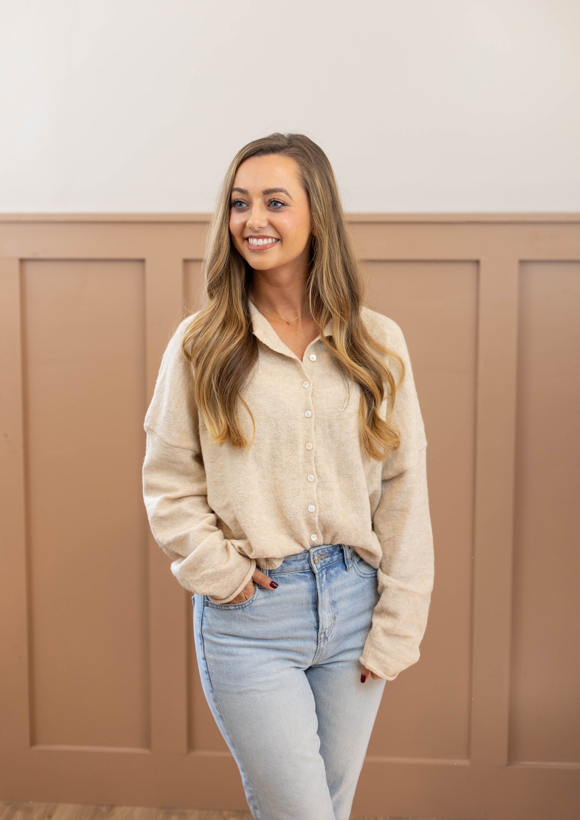 A woman with long hair, wearing the cozy Clay Button-Down Sweater in an oatmeal hue from Dogwood Cloth and blue jeans, smiles while standing in front of a paneled wall.