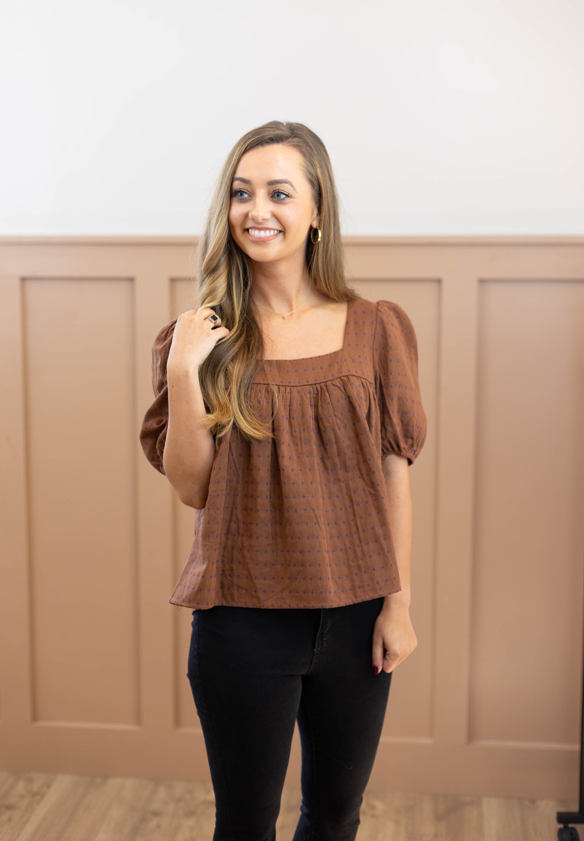 A woman with long hair is smiling and looking to the side while standing in front of a light brown paneled wall. She is wearing the Cara Timeless Top by Dogwood Cloth, which has a subtle embroidered pattern that complements its square neckline, along with black pants.