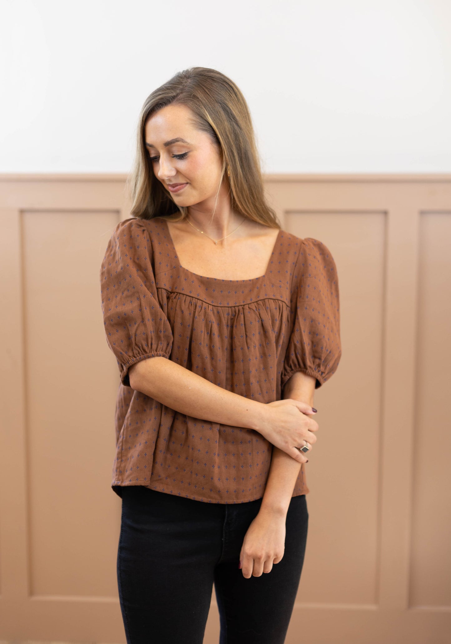 A person stands against a paneled wall, looking down. They wear the Cara Timeless Top from Dogwood Cloth, featuring puffed sleeves and a delicate embroidered pattern, complemented by black pants.