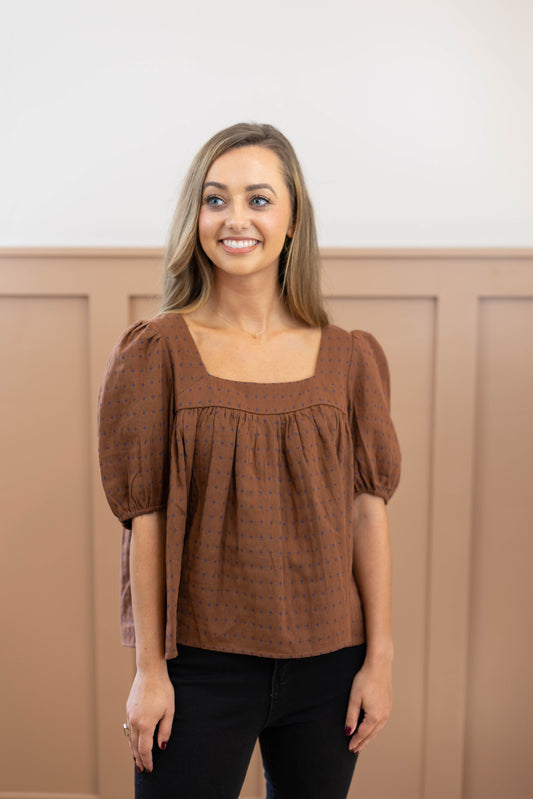 A person with long hair is wearing a Cara Timeless Top from Dogwood Cloth in brown, along with black pants, as they stand and smile slightly in front of a beige paneled wall.