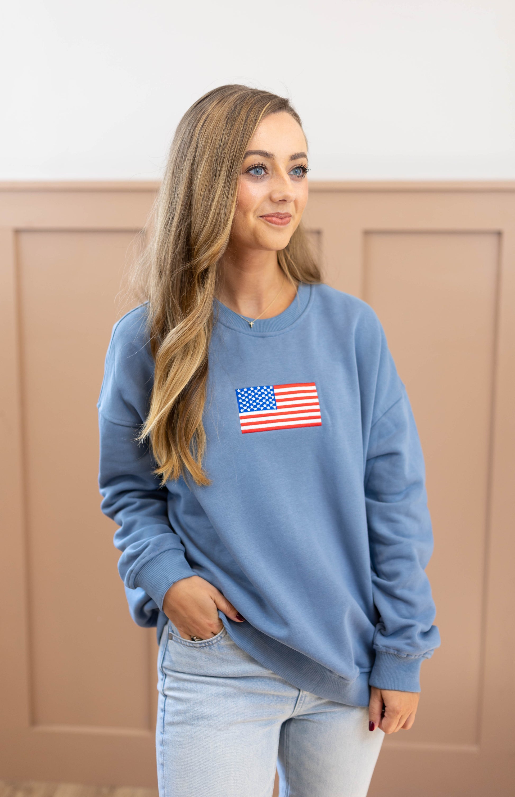 A person with long hair is wearing a denim blue Liberty Crewneck Pullover by Dogwood Cloth, featuring a small American flag patch, standing against a light brown paneled wall.