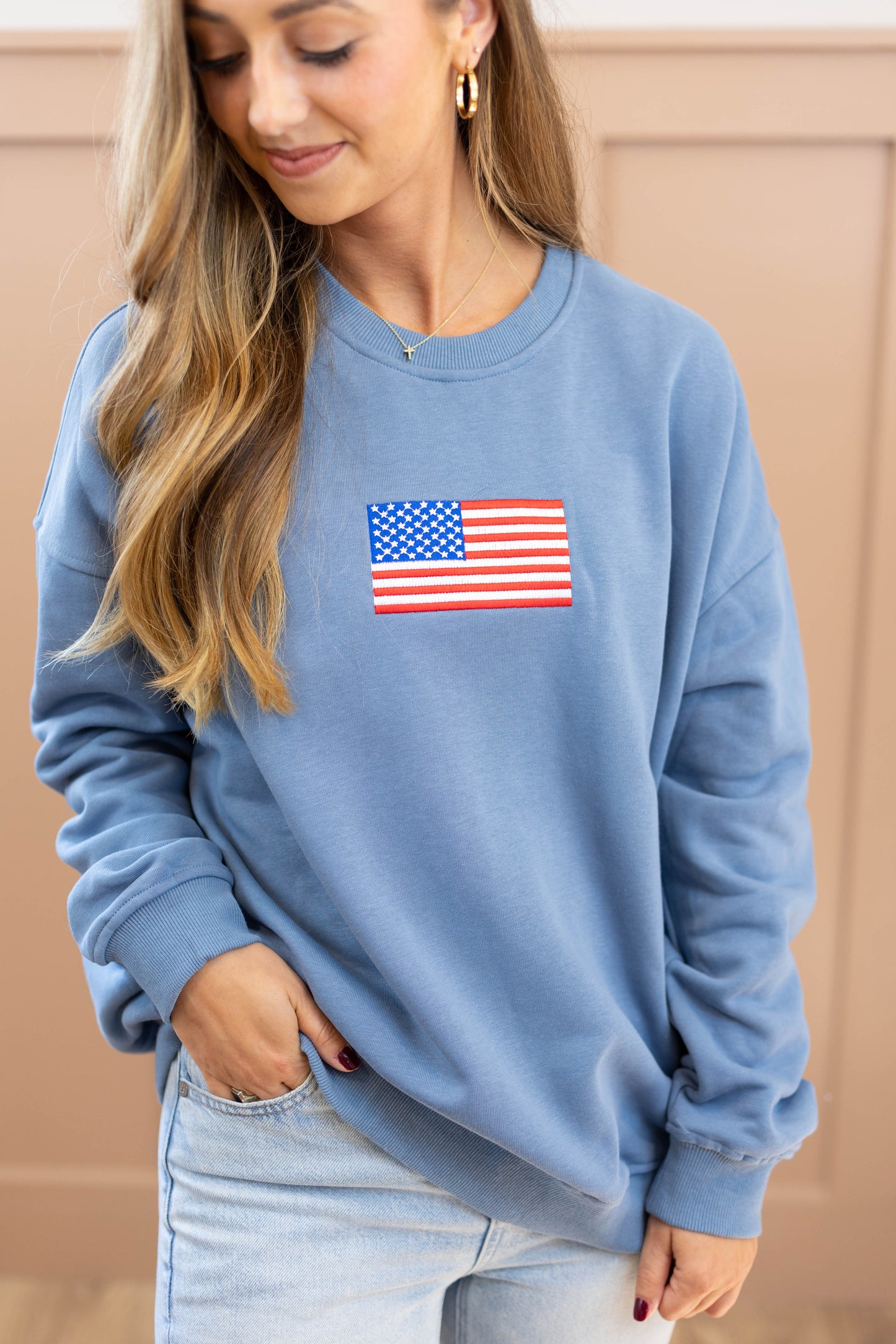 A person wearing the Dogwood Cloth Liberty Crewneck Pullover, featuring an American flag patch on the chest, stands against a beige background.