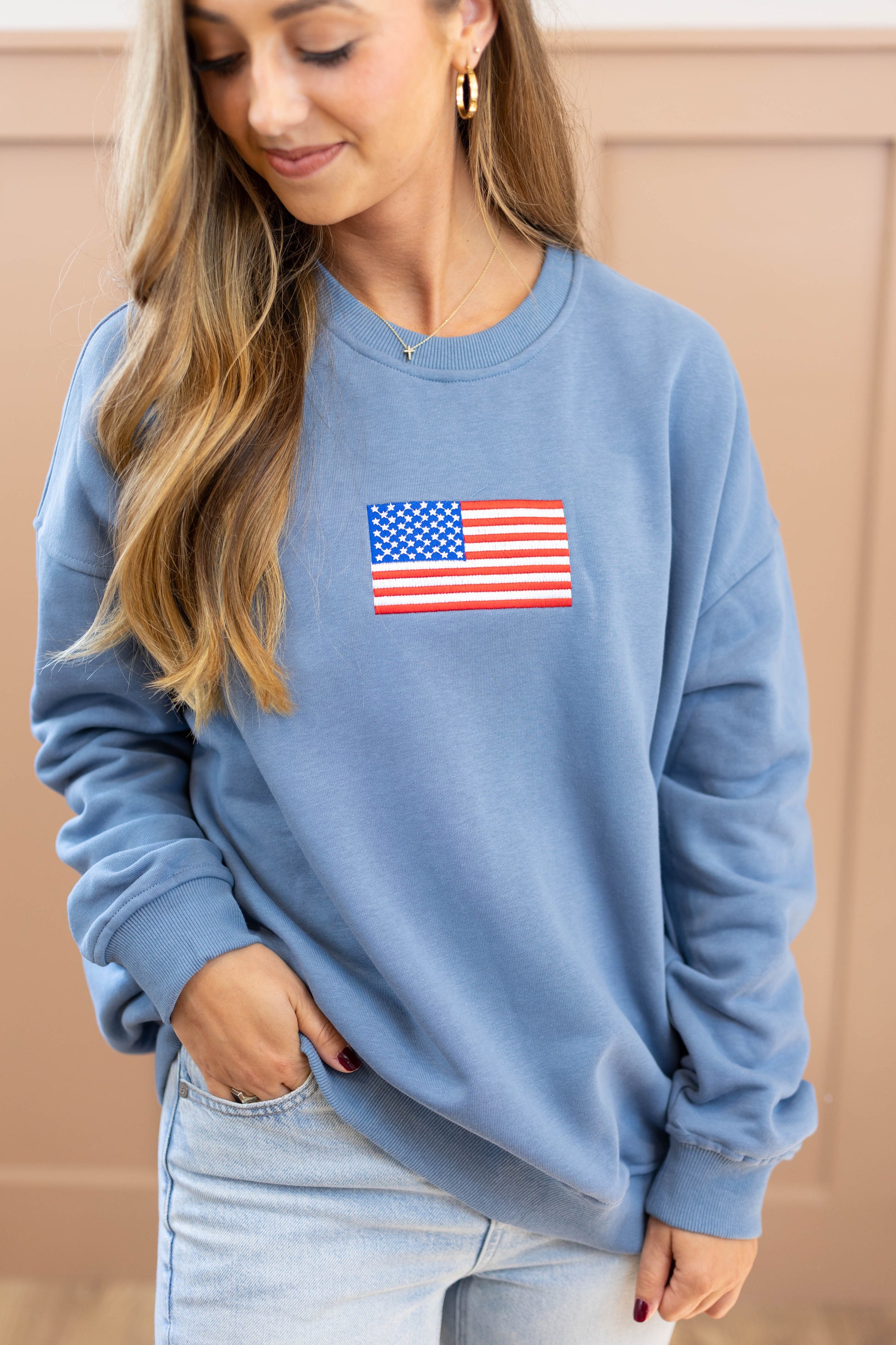 A person wearing the Dogwood Cloth Liberty Crewneck Pullover, featuring an American flag patch on the chest, stands against a beige background.