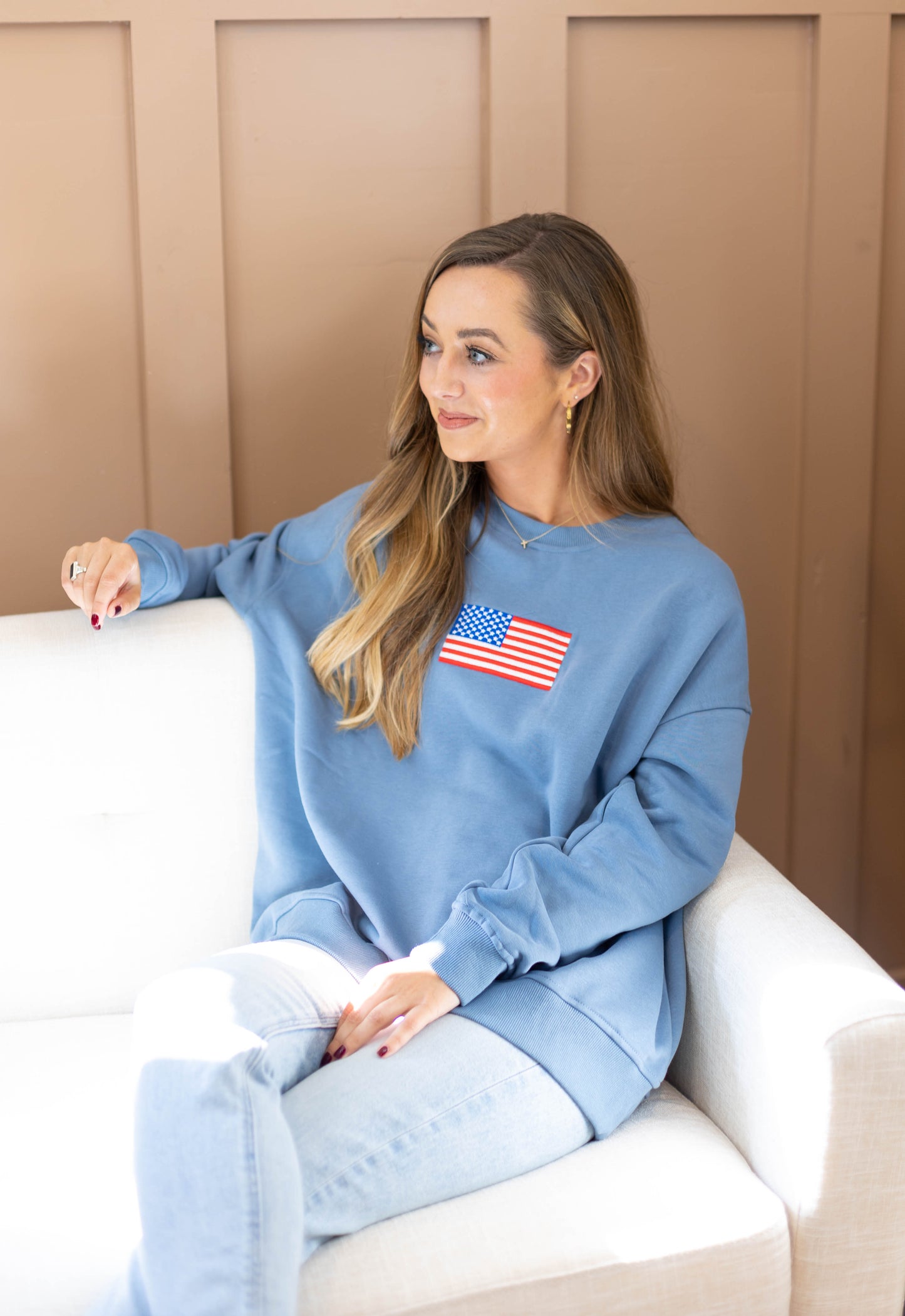 A person wearing an ultra-soft Liberty Crewneck Pullover by Dogwood Cloth, featuring an American flag patch, sits on a white couch against a tan wall.