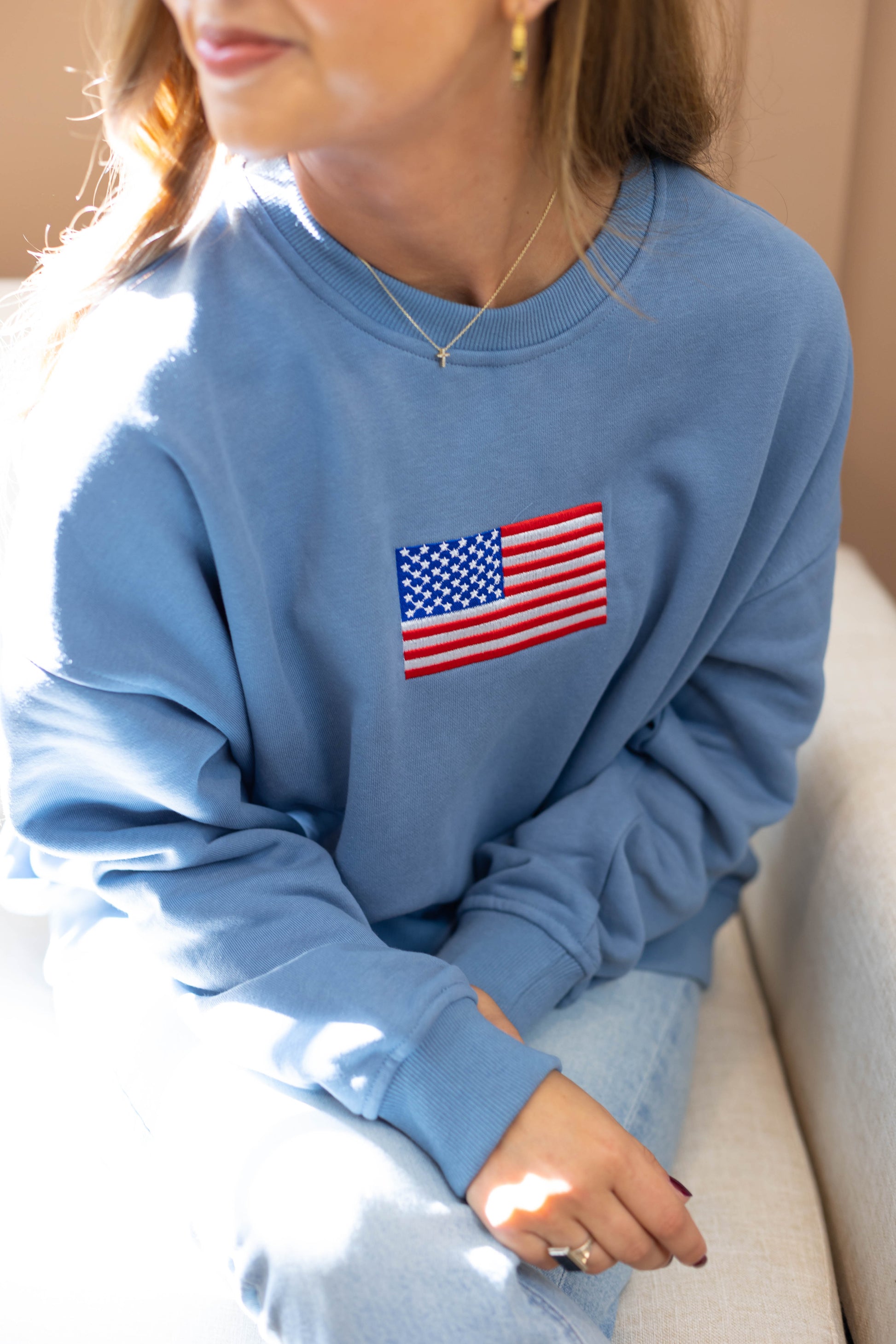 A person is cozily seated on a light-colored couch, wearing an ultra-soft Liberty Crewneck Pullover by Dogwood Cloth, featuring an embroidered American flag design.