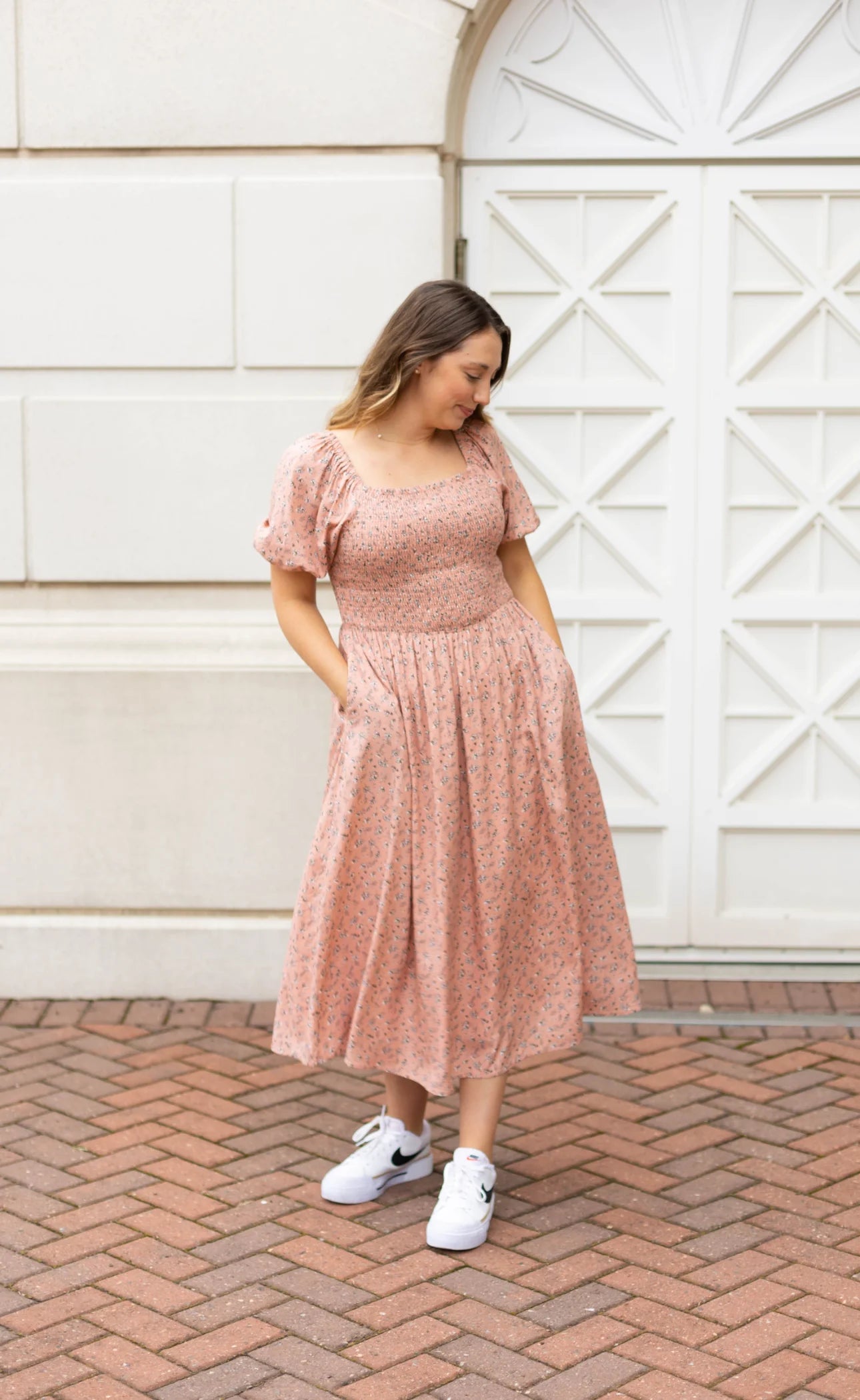 A woman wearing the Blakely Midi Dress by Dogwood Cloth, featuring a blush-rose floral pattern with a smocked bodice and pockets, pairs it with white sneakers while standing on a brick pathway in front of a white door.