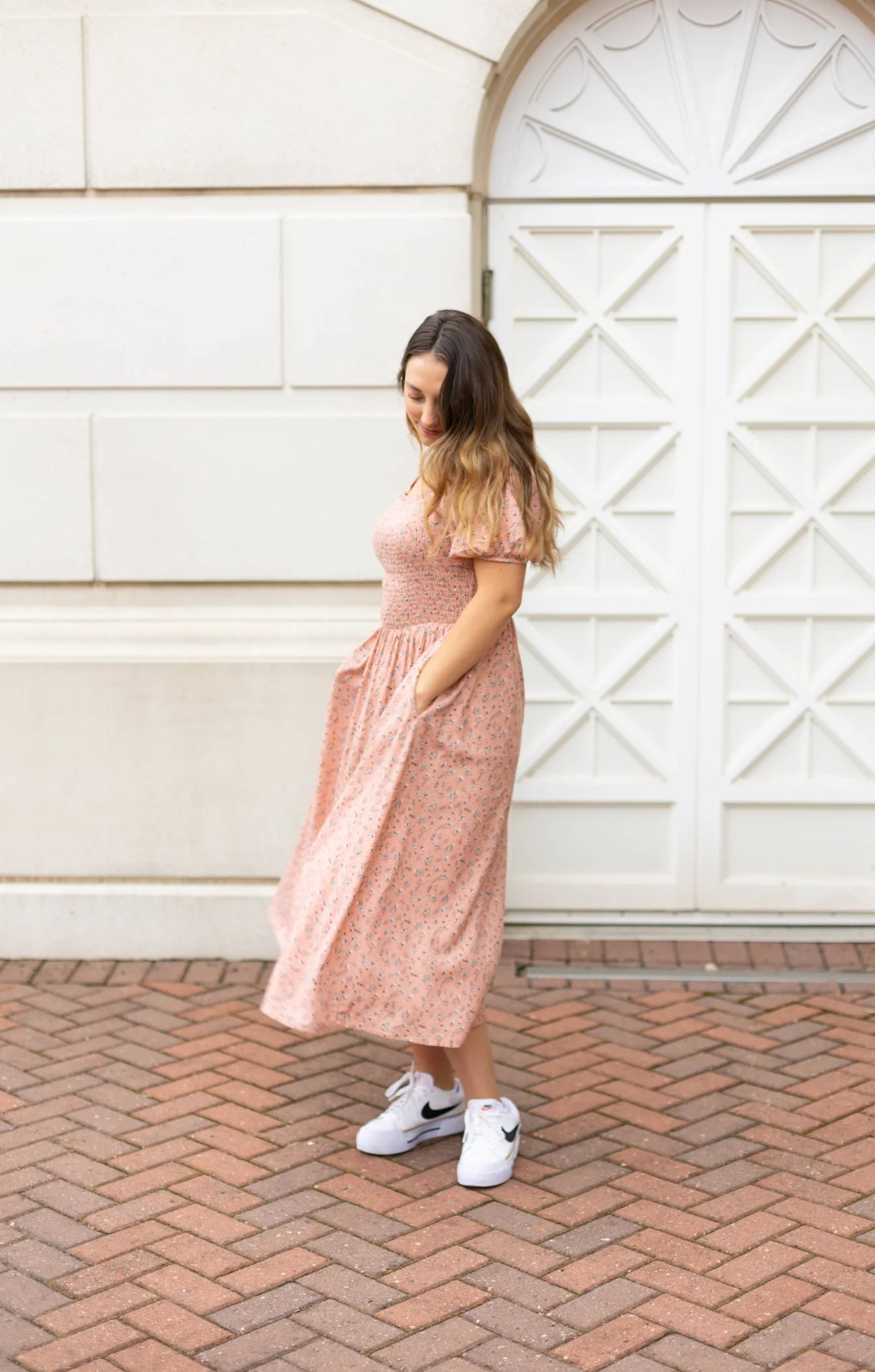 A woman wearing a Blakely Midi Dress from Dogwood Cloth, featuring a blush-rose floral pattern, pairs it with white sneakers as she stands on a brick path in front of a geometric white door.