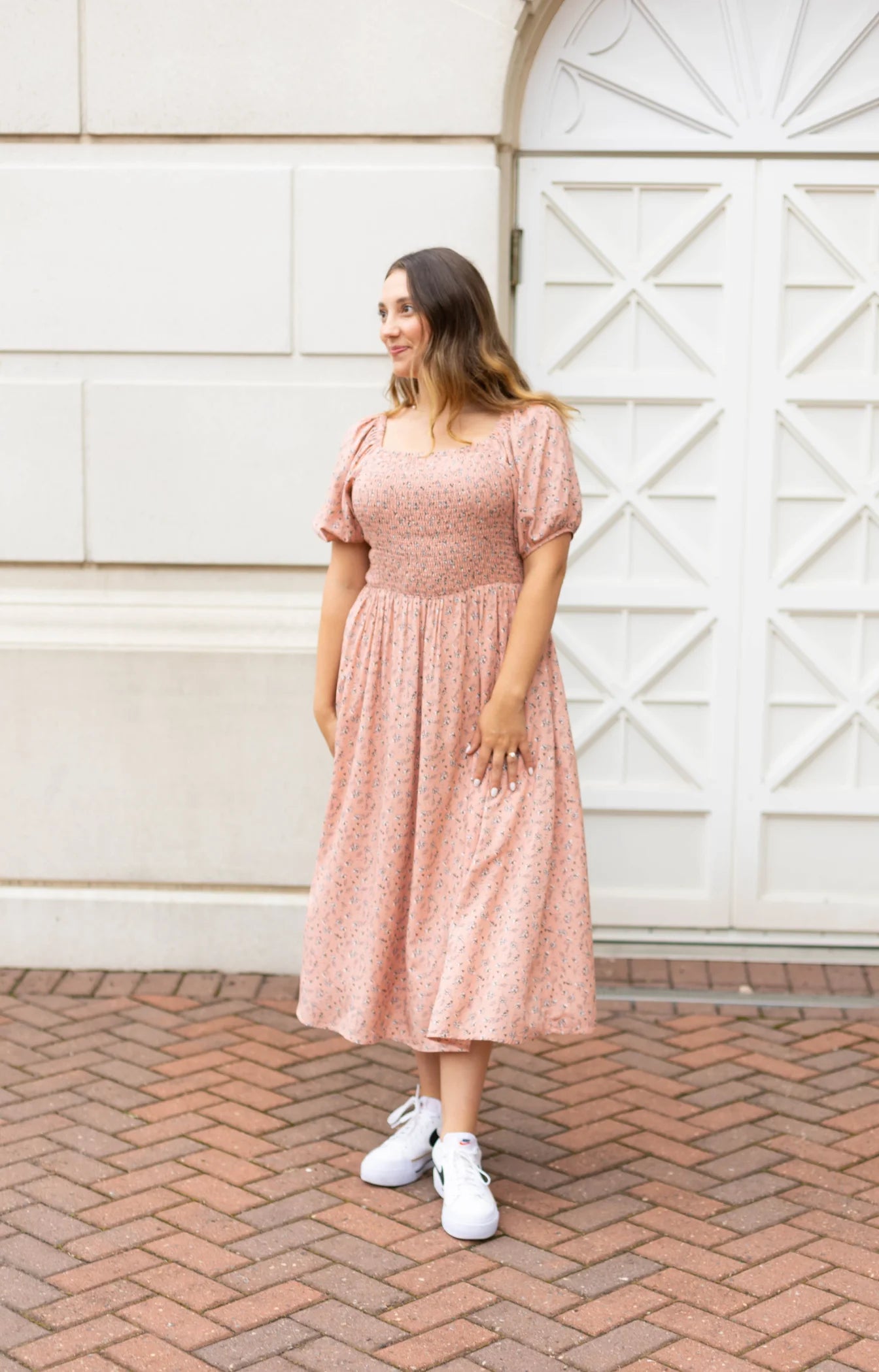 A person in a Dogwood Cloth Blakely Midi Dress featuring a blush-rose floral design and wearing white sneakers stands on a brick walkway in front of an ornate white door.