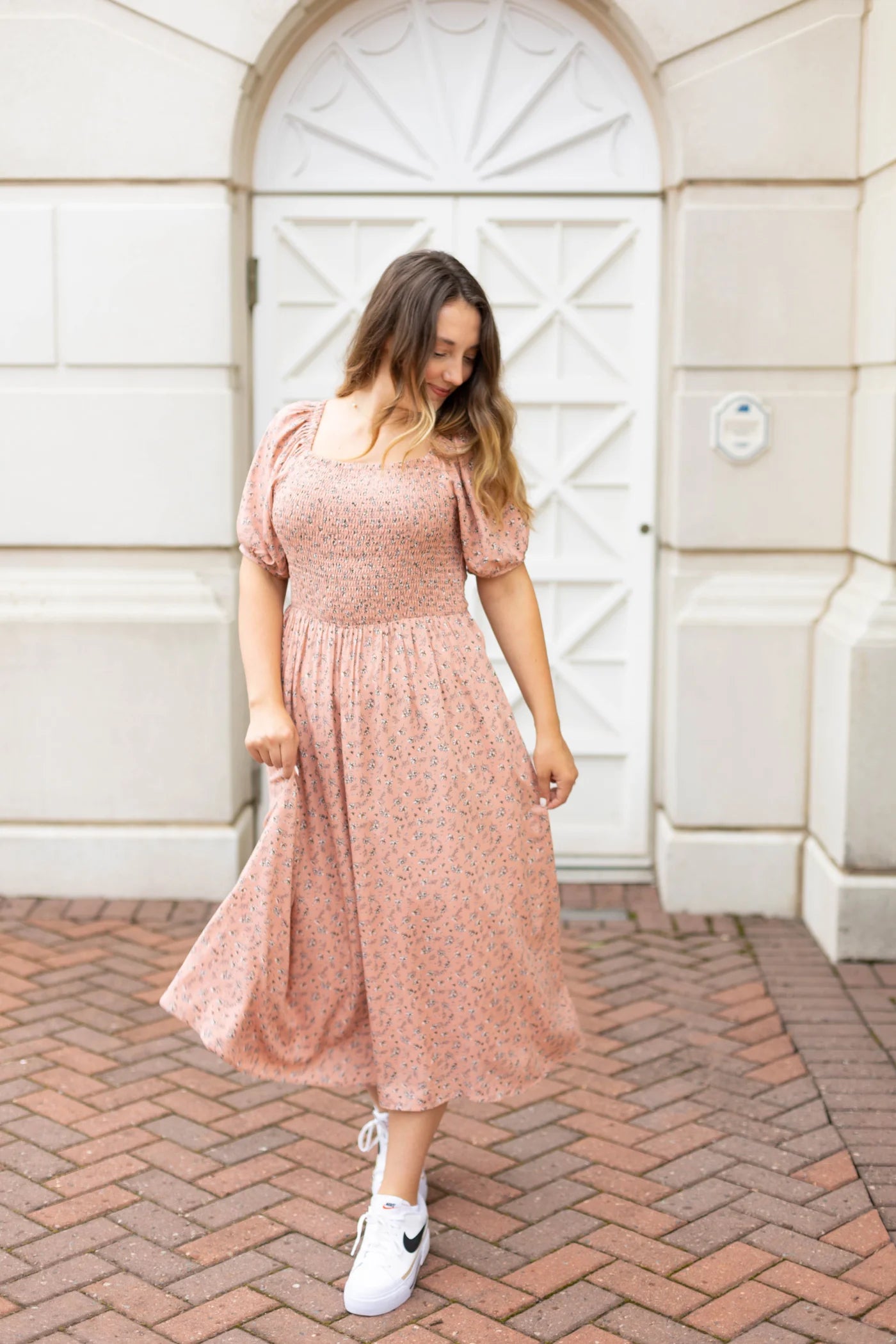 A person in a Blakely Midi Dress by Dogwood Cloth and white sneakers stands on a brick path in front of an arched white door.