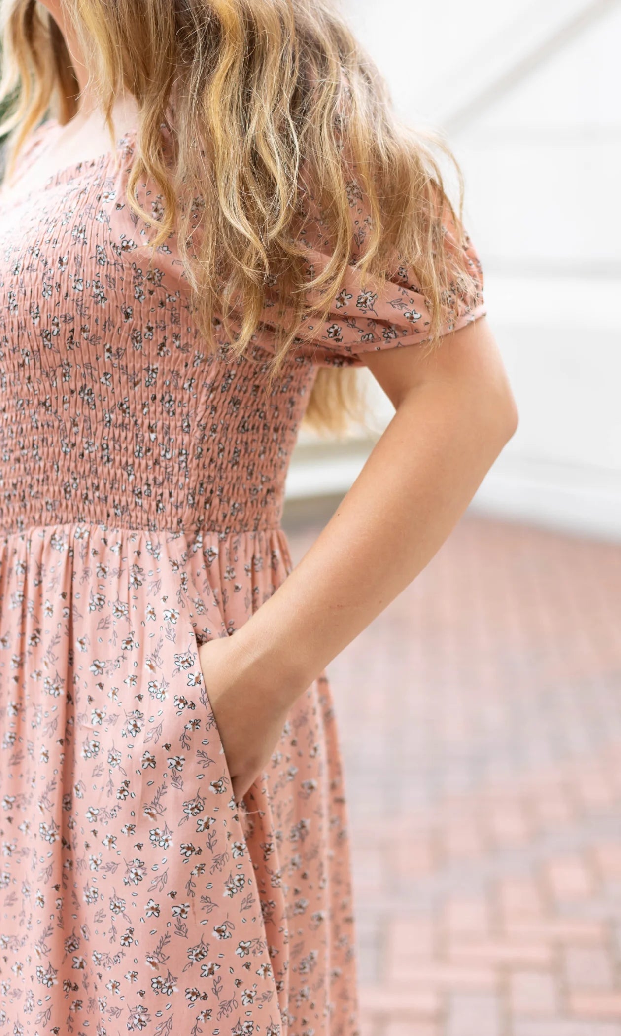 A person stands outdoors, their hand casually placed in the side pocket of a Dogwood Cloth Blakely Midi Dress. The dress showcases a smocked bodice with a blush-rose floral pattern, adding an elegant touch to their look.