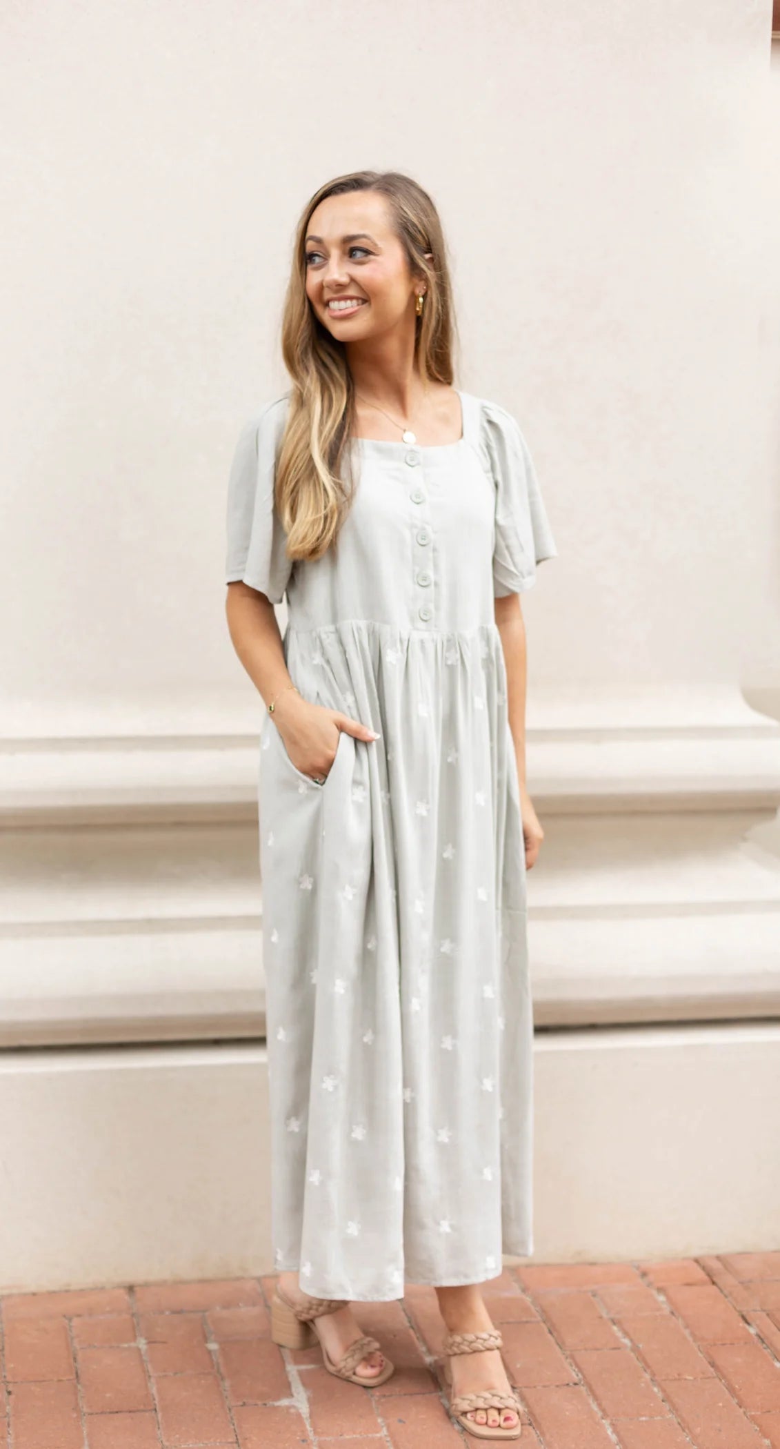 A person wearing a Lydia Embroidered Midi Dress by Dogwood Cloth, featuring a light gray hue with buttoned front, flutter sleeves, and embroidered floral details, stands against a light-colored wall, smiling and gazing to the side.