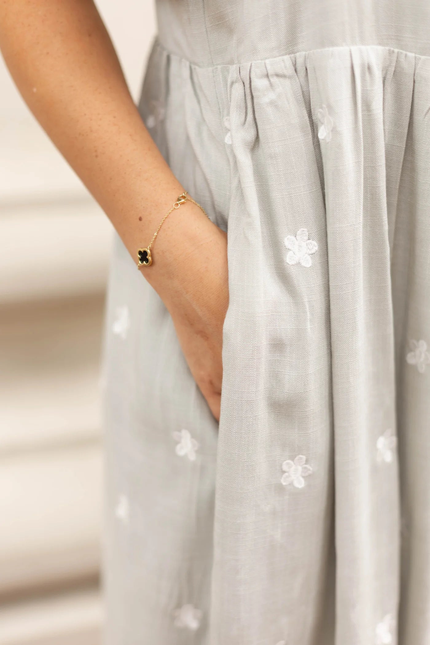 A person dressed in a light gray Lydia Embroidered Midi Dress by Dogwood Cloth, detailed with white embroidered flowers and flutter sleeves, casually places a hand in the dress pocket, showcasing a gold bracelet adorned with a four-leaf clover charm.